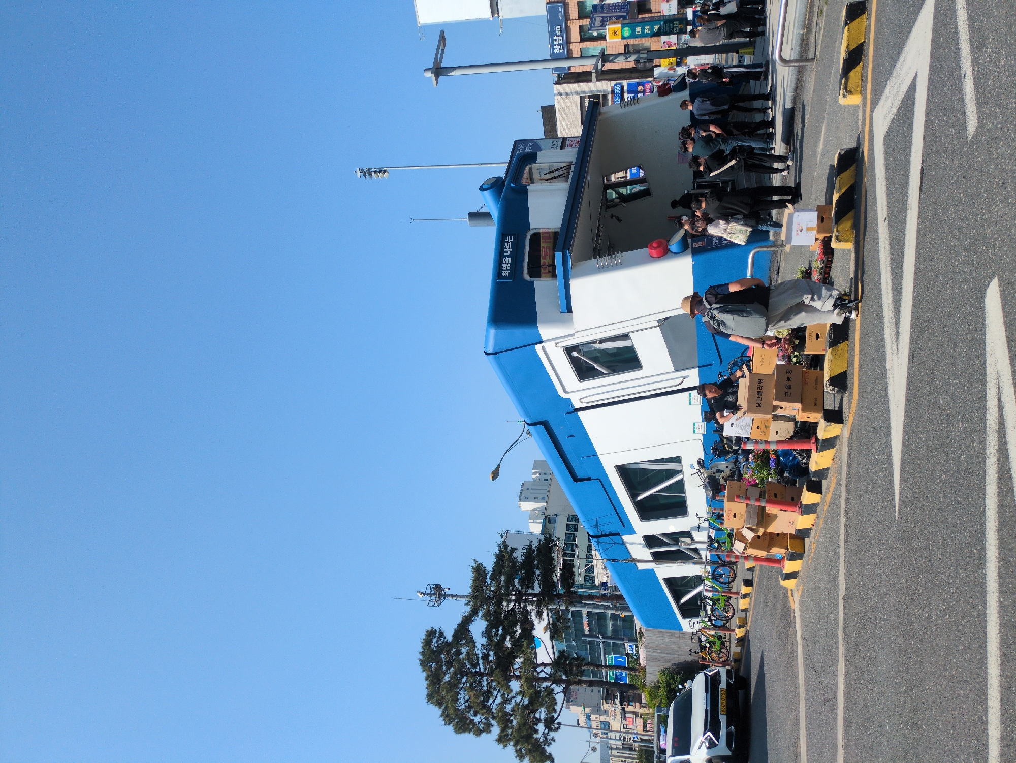 The image depicts a train-shaped structure in the city of Daejeon, South Korea. The structure, painted in blue and white, is identified by signage in Korean as "낭만폴라워" (Romance Flower) and "근육농장" (Muscle Farm), and a phrase, "희망을 나르는" (Carrying Hope). People are seen inside the structure. Boxes are situated next to the vehicle. Bicycles are parked nearby. A white van is also parked on the side of the road. A road marked with yellow and black posts lies adjacent to the structure. In the background, buildings and a clear blue sky are visible.