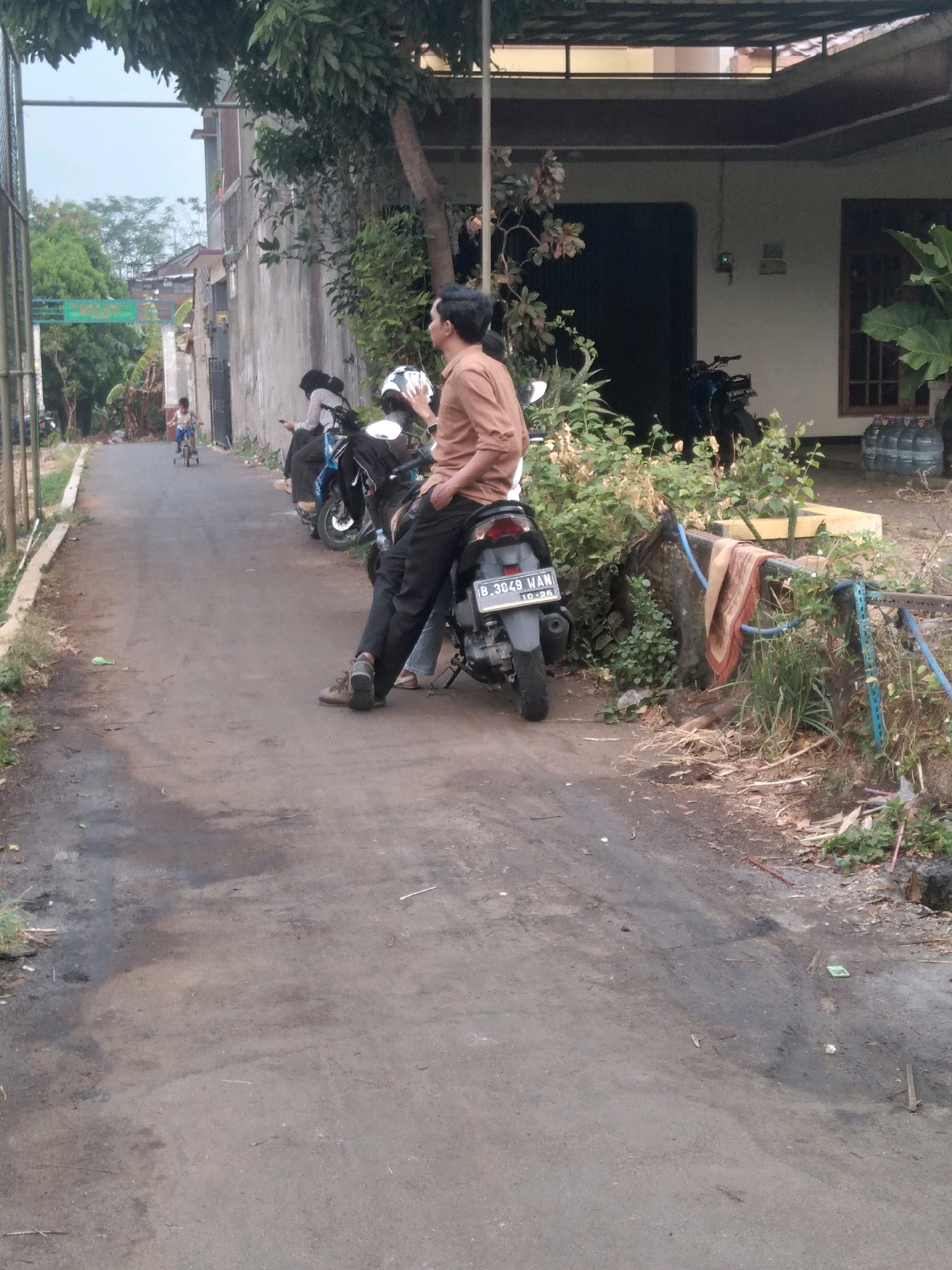 In South Tangerang, Indonesia, a dirt road curves away from the camera, bordered by a rough concrete wall on the left and overgrown vegetation and buildings on the right. Two men are the primary subjects; one, wearing a brown shirt and black pants, sits casually on a motorcycle parked on the side of the road, facing away from the camera with his right hand in his pocket. Another person, partially obscured by the first, sits on the same motorcycle. To their left, another motorcycle is parked with two individuals, one wearing a black headscarf and a striped shirt. Further down the road, a young child rides a bicycle away from the camera. The environment appears somewhat unkempt, with debris scattered on the road and vegetation encroaching on the edges. A building with dark garage doors and a tiled roof is visible in the background. A rack with a colorful rug draped over it is positioned among the plants on the right side of the road.