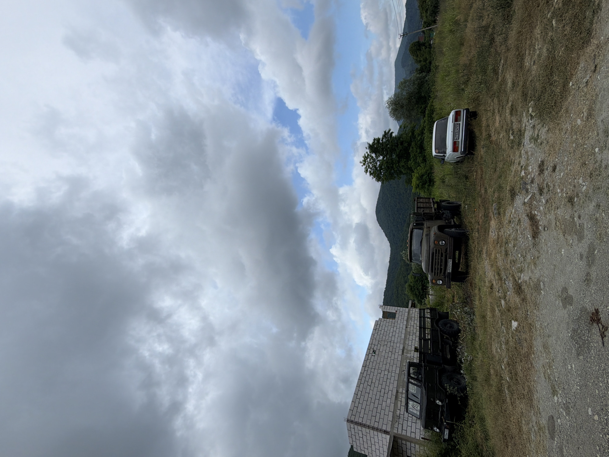 In Shepsi, Russia, under a dramatic sky filled with heavy, grey clouds and patches of blue, a construction site features an unfinished building made of white brick blocks. To the left of the building, a dark green, older model truck is parked on a grassy area. Further to the right, a khaki-colored truck, also an older model, sits in the tall grass, its headlights visible. Beside it, a white sedan is parked. In the background, lush green, tree-covered mountains rise against the cloudy sky. The foreground consists of a gravelly area.