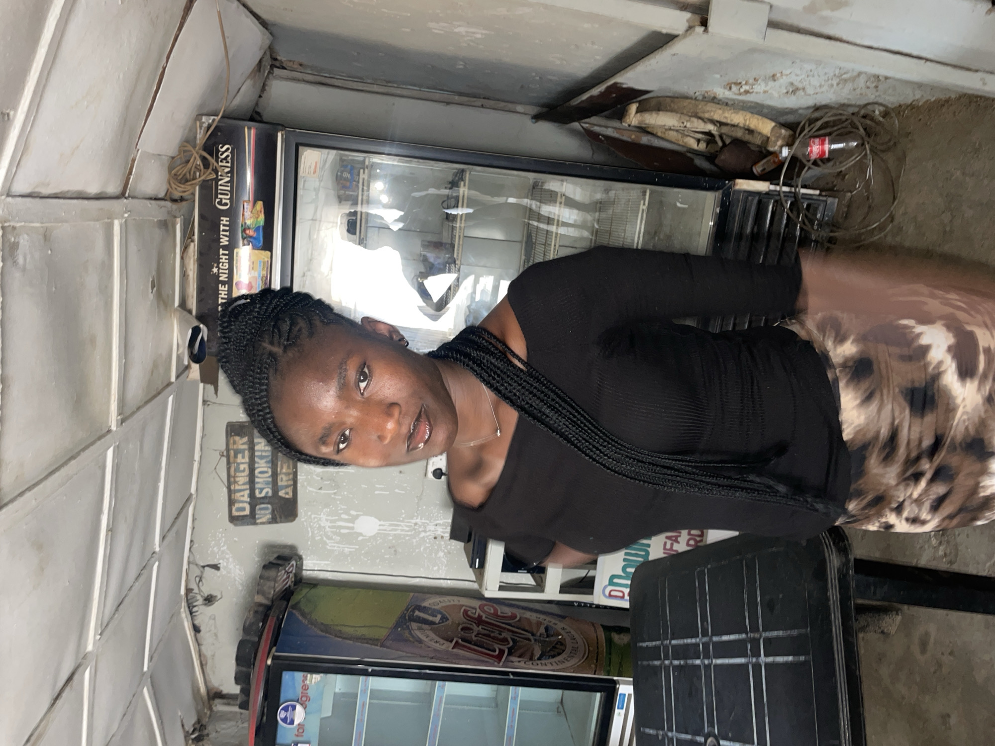 A young woman with dark skin and braided hair stands posing for a photo in what appears to be a small, utilitarian shop or kiosk. She wears a black ribbed top and a brown and tan patterned skirt or lower garment. A simple necklace adorns her neck, and she looks directly at the camera with a neutral to slightly smiling expression.
The setting is an indoor or semi-enclosed space, likely a commercial establishment, possibly in Jos, Nigeria. The walls are constructed from light-colored, possibly corrugated metal or painted panels, showing signs of wear, discoloration, and minor damage. The floor is concrete.
Behind the woman are two commercial display refrigerators or coolers. The one to her left features a prominent "GUINNESS" logo and the text "THE NIGHT WITH GUINNESS." The other refrigerator, partially visible to her right, displays a large "Lite" beer logo, characteristic of Miller Lite. Between the two refrigerators, in the upper background, is a collection of various wires, including a coiled bundle, and a red Coca-Cola bottle. Above a lower refrigerator on the left, a sign reads "DANGER NO SMOKING AREA." Other labels on the coolers are partially visible, including one with the text "for gress." A black plastic chair or stool is visible in the foreground to the right. The overall scene suggests a modest local business.