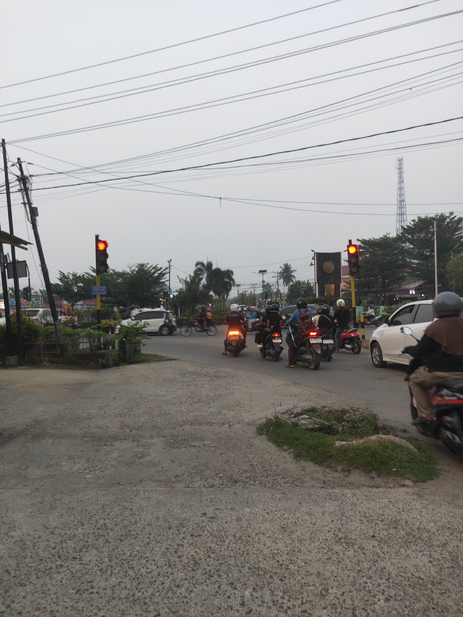 This is an outdoor, medium-shot, eye-level photograph taken in Sintang, Indonesia, likely during the late afternoon or early evening, as indicated by the overcast sky and traffic light signals. The scene depicts a busy intersection with several motorbikes and a few cars.
In the foreground, the road surface is cracked and shows signs of wear, with patches of grass growing in a depression on the right side. A motorcycle with a rider wearing a helmet and a brown headscarf is visible on the right, moving away from the camera.
Further into the scene, a traffic light with a red light illuminated stands prominently on a yellow pole. A white car is stopped near the traffic light, and several motorbikes are lined up, waiting for the signal to change. Some riders are wearing helmets, while others are not. The general atmosphere suggests a typical urban traffic situation.
In the background, more trees and buildings can be seen, along with power lines crisscrossing the sky. A tall telecommunications tower is visible in the far distance. The overall environment appears to be a moderately developed urban area.