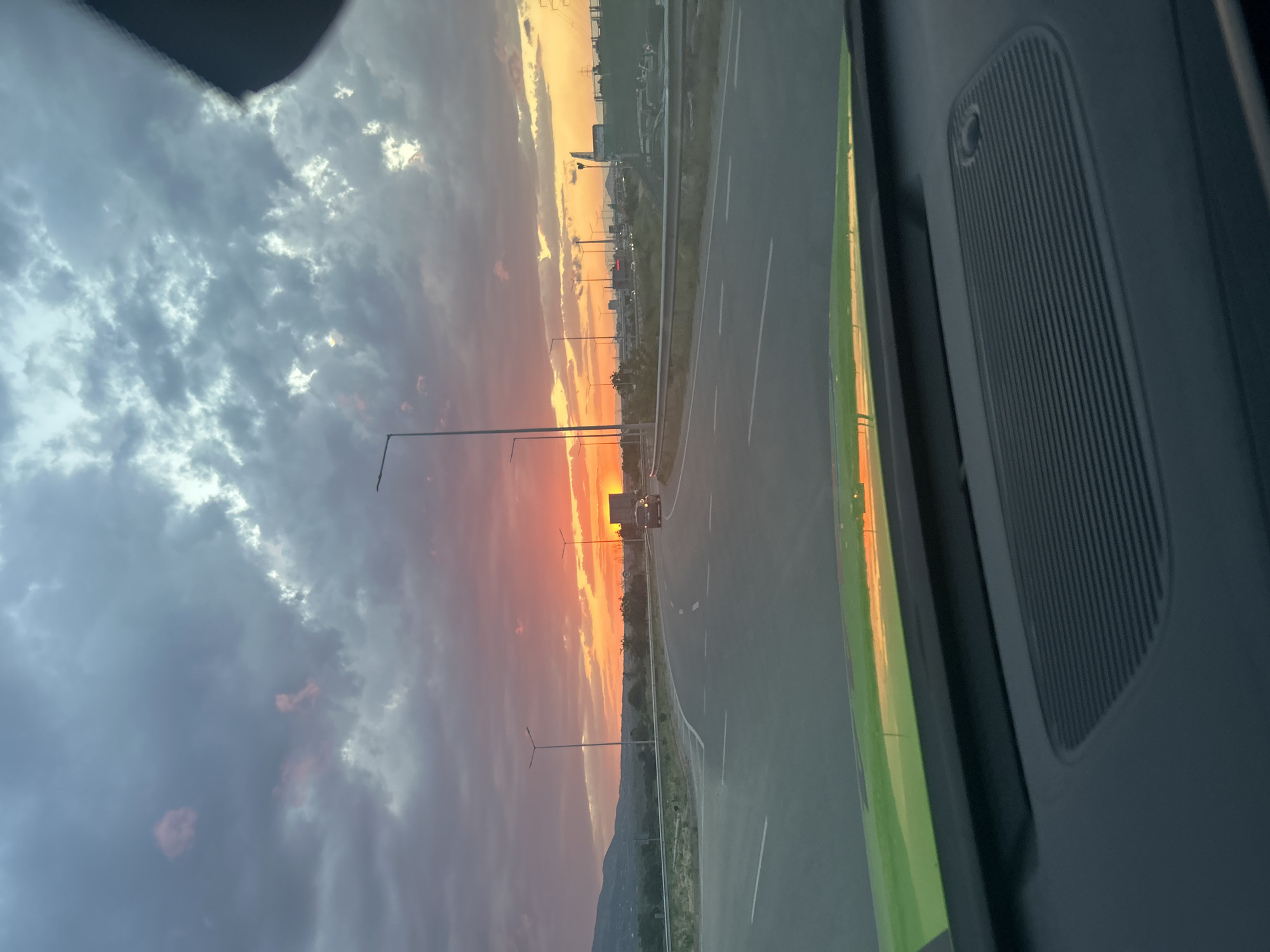 Here's a detailed description of the image:
**Scene:** The image captures a view from inside a vehicle, likely a car, looking out through the side window. A section of the vehicle's interior, including a speaker grill, is visible on the right side of the frame.
**Environment:** The exterior scene depicts a road, likely a highway or major thoroughfare, with multiple lanes. Streetlights are present along the roadside. The sky is filled with clouds, with a striking sunset occurring in the distance. The sunset displays colors of orange, pink, and yellow blending with the gray and blue of the clouds. A mountain range or large hills are visible on the horizon.
**Location:** Unknown City, Unknown. The details are not resolvable.