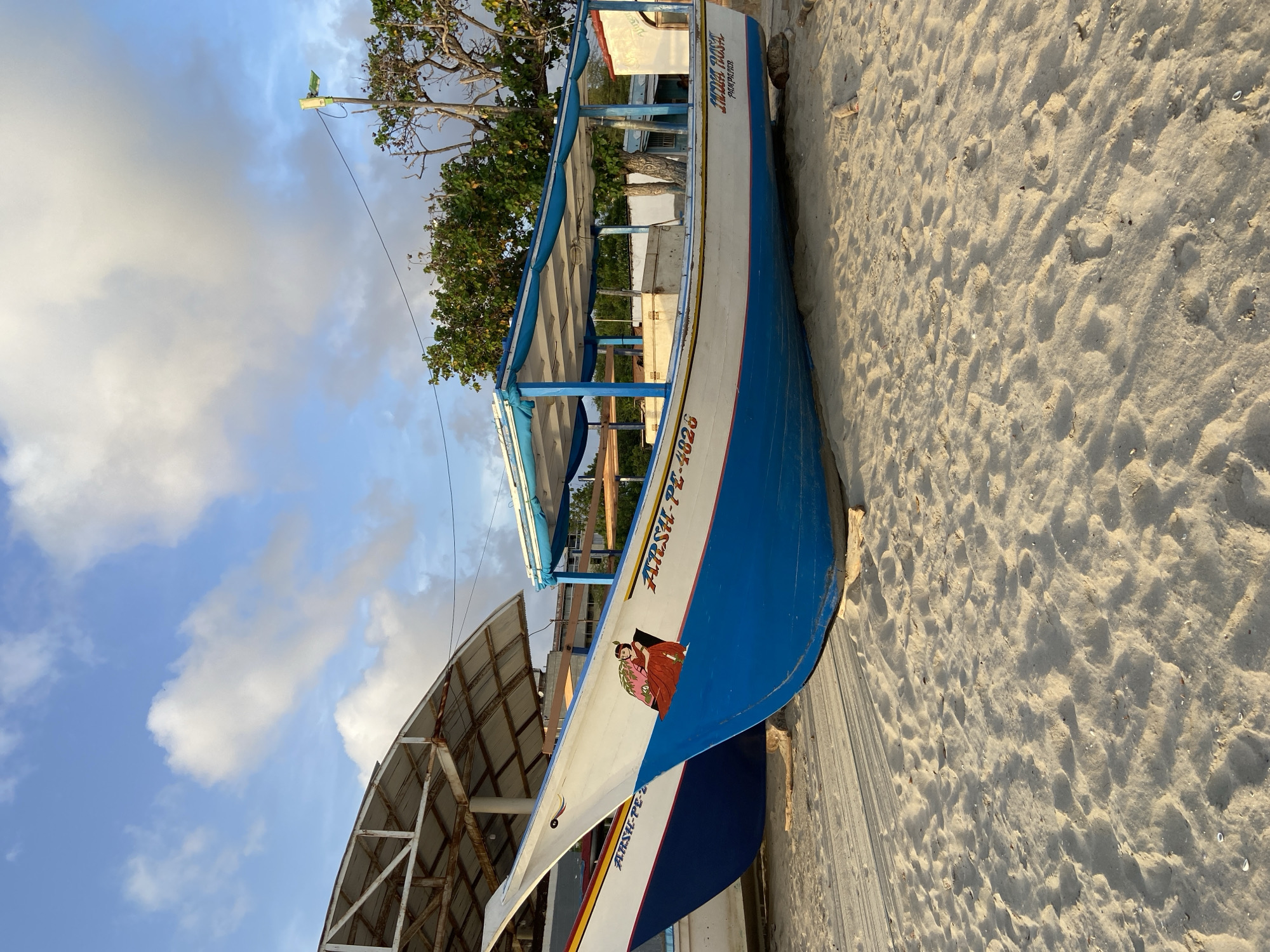The image depicts several boats resting on a sandy beach in Juan Griego, Venezuela, under a partly cloudy sky. The boats are colorful, with white and blue exteriors, and some are decorated with painted imagery and text. One prominent boat in the foreground has the inscription "ARSHI-PE-2828" and a depiction of a woman in traditional attire. Another boat behind it also has similar markings. The beach is composed of light-colored sand with scattered debris. In the background, a structure with a corrugated metal roof, possibly a covered market or storage area, is visible, along with trees and a distant building. The lighting suggests it is either early morning or late afternoon.
