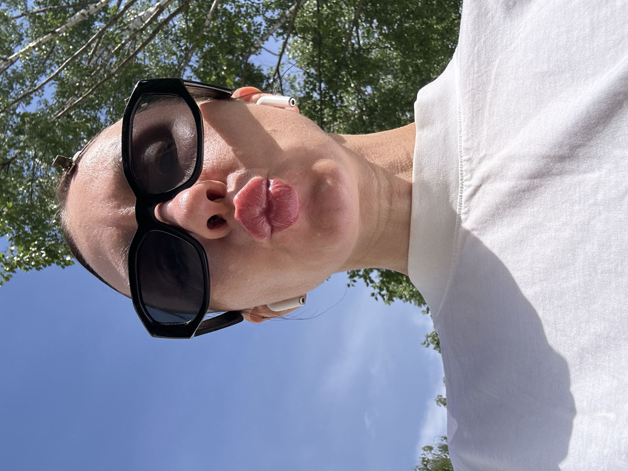 The image shows a person with fair skin, wearing black sunglasses and white wireless earbuds. The person is making a kissing face. They are wearing a white shirt. The background includes blue sky and green foliage, possibly from trees. The location is not identifiable as Votkinsk, Russia based on the information presented in the image.