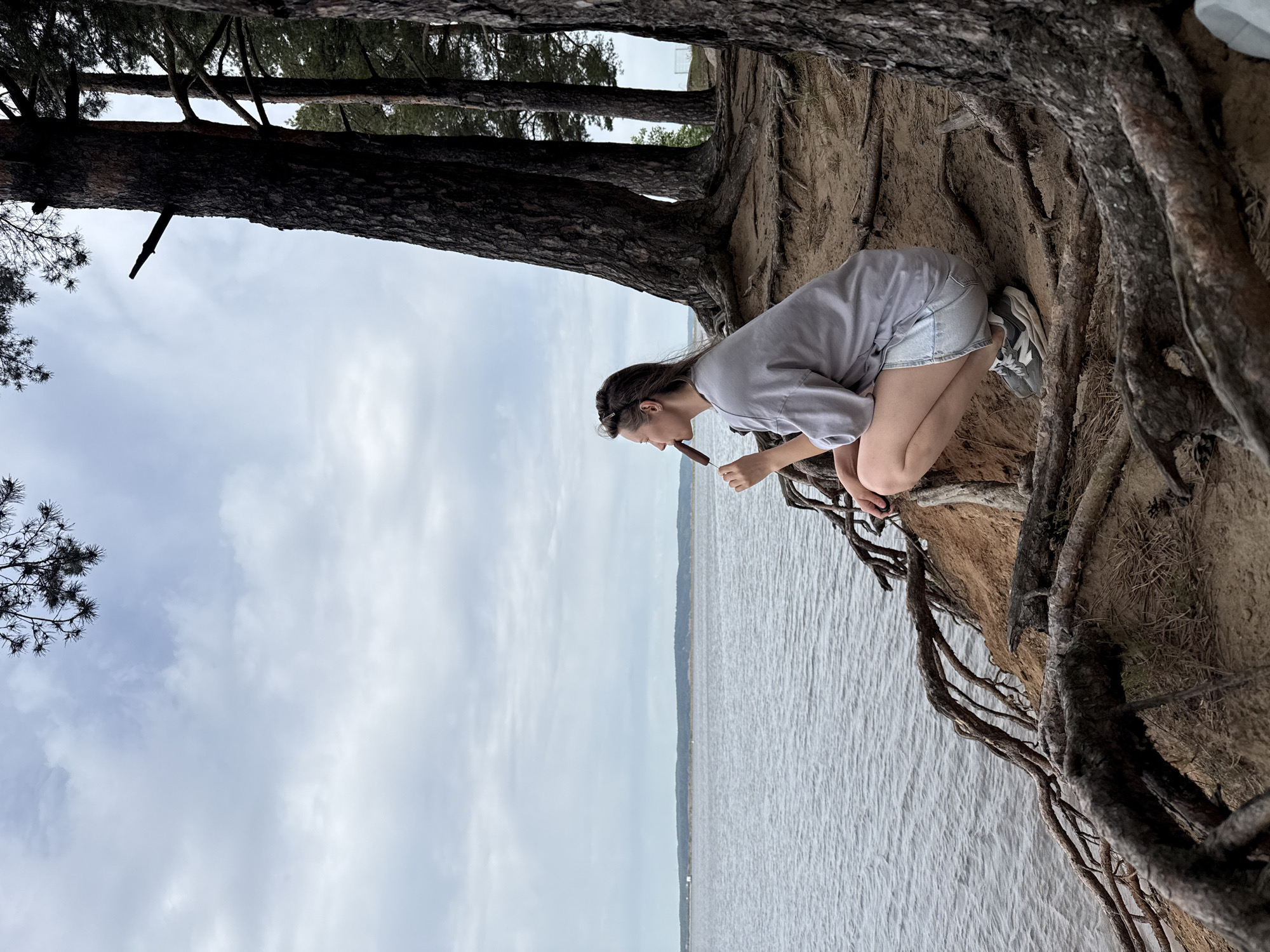 A woman is kneeling on a sandy embankment at a waterside location in the city of Chaikovsky, Russia. She is holding a chocolate-covered ice cream bar in her hand, seemingly about to take a bite. The embankment is covered with trees and exposed roots. The water and sky dominate the background.