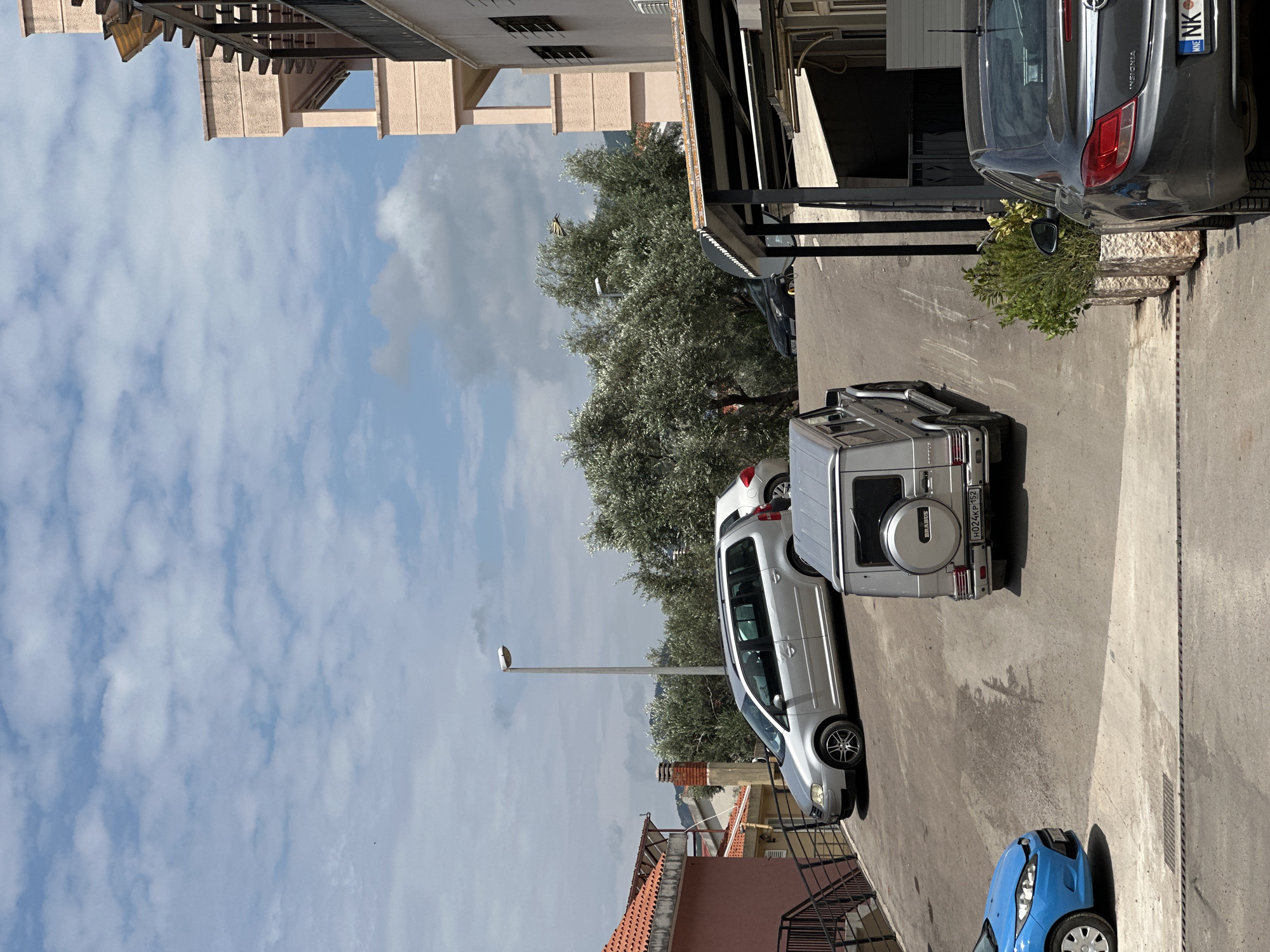 The image depicts a bright, daytime scene of a street or parking area in what appears to be a residential or urban setting. The sky, partially covered by white cumulus clouds, dominates the upper portion. Below the sky, several multi-story buildings with light-colored facades and balconies or pergolas are visible. Numerous olive trees with their characteristic grey-green foliage line one side of the street.
Several vehicles are parked on a paved surface. In the foreground, a silver Mercedes G-Wagen SUV, identified by the "BRABUS" text on its rear spare tire cover and license plate "H024KP 152", is prominently featured. Next to it is a silver minivan or station wagon. Further down the street, a dark grey Opel Insignia sedan is parked, displaying "INSIGNIA" on its trunk and a license plate with "NK," indicating Nikšić, Montenegro. A small blue car is partially visible in the lower right foreground. Another dark vehicle is parked under an overhang.
A single person is partially visible among the olive trees. A large, yellow butterfly, likely a swallowtail, is perched on one of the olive trees. A street light pole stands near the trees. The strong shadows suggest it is mid-day. The overall scene is calm and portrays a typical street view.