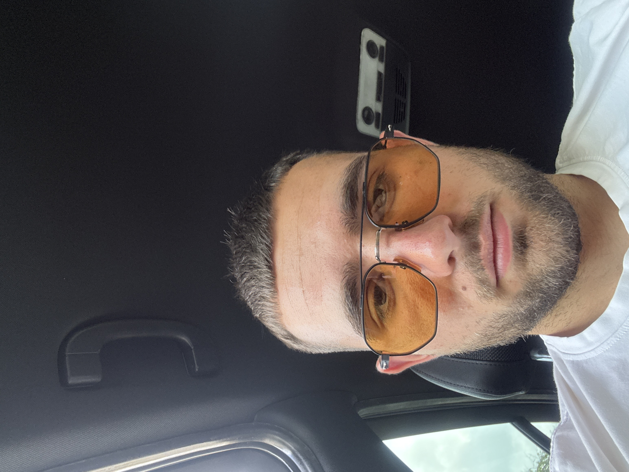 A man is looking at the camera, wearing orange tinted glasses and a white shirt in a car. The interior of the car has a black ceiling. There is a handle above the window on the left and a light fixture on the right. The image shows a partial view outside of the car. There is no information to connect the image to Gabrovo, Bulgaria.