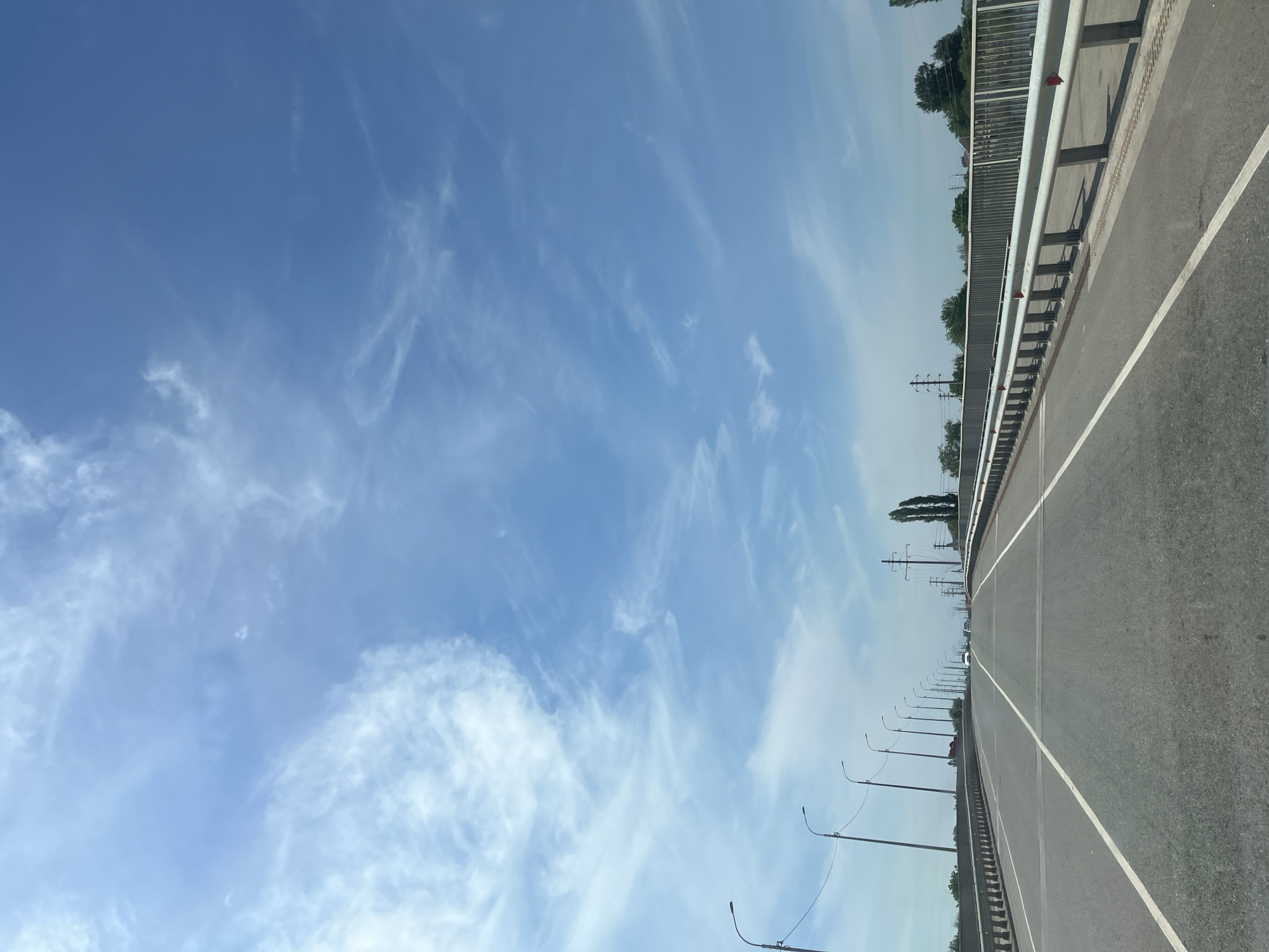 The image shows a view from a car traveling on a road, likely a bridge, in Krasnyye Barrikady, Russia. The road is paved with asphalt and has white lane markings. On the left side of the road, there are several streetlights. On the right side, there is a metal guardrail with red reflectors. In the distance, there are trees and some power lines. The sky is blue with scattered white clouds. There are no people visible in the image, and no actions or interactions are depicted. The environment appears to be a typical urban or suburban setting with infrastructure for transportation.