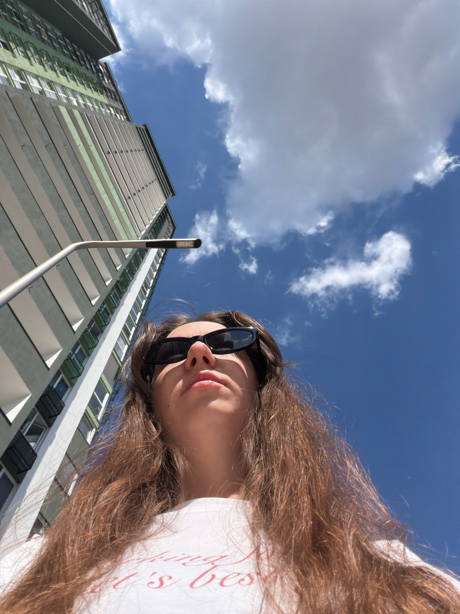 A low-angle, close-up, outdoor, daytime, eye-level shot shows the top of a young person's head and shoulders, looking upwards. The person has long, brown hair, is wearing black sunglasses, and a white t-shirt with pink text. Behind them, a modern, tall apartment building with a green and gray facade is visible, along with a street lamp. The sky is bright blue with scattered white clouds. The image is likely taken in Kyiv, Ukraine, given the architectural style of the building and common weather conditions for the region.