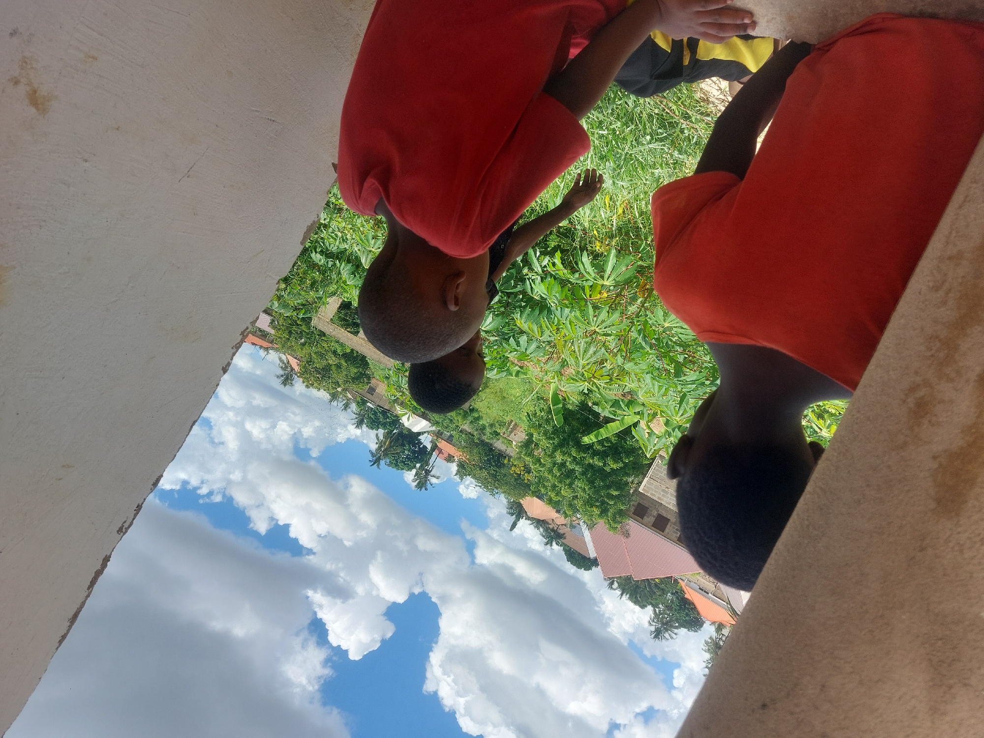 Two children are seen in an outdoor setting. Both are wearing orange shirts. One child is seen partially, with their head and torso visible, while the other is visible from the head down to the upper thighs. There appears to be green vegetation in the area. A building is visible in the background. The sky is visible in the upper right. The children are interacting with something in the foliage, and the photo is taken from the perspective of someone under an overhang or doorway. The building and vegetation suggest Dar es Salaam, Tanzania.