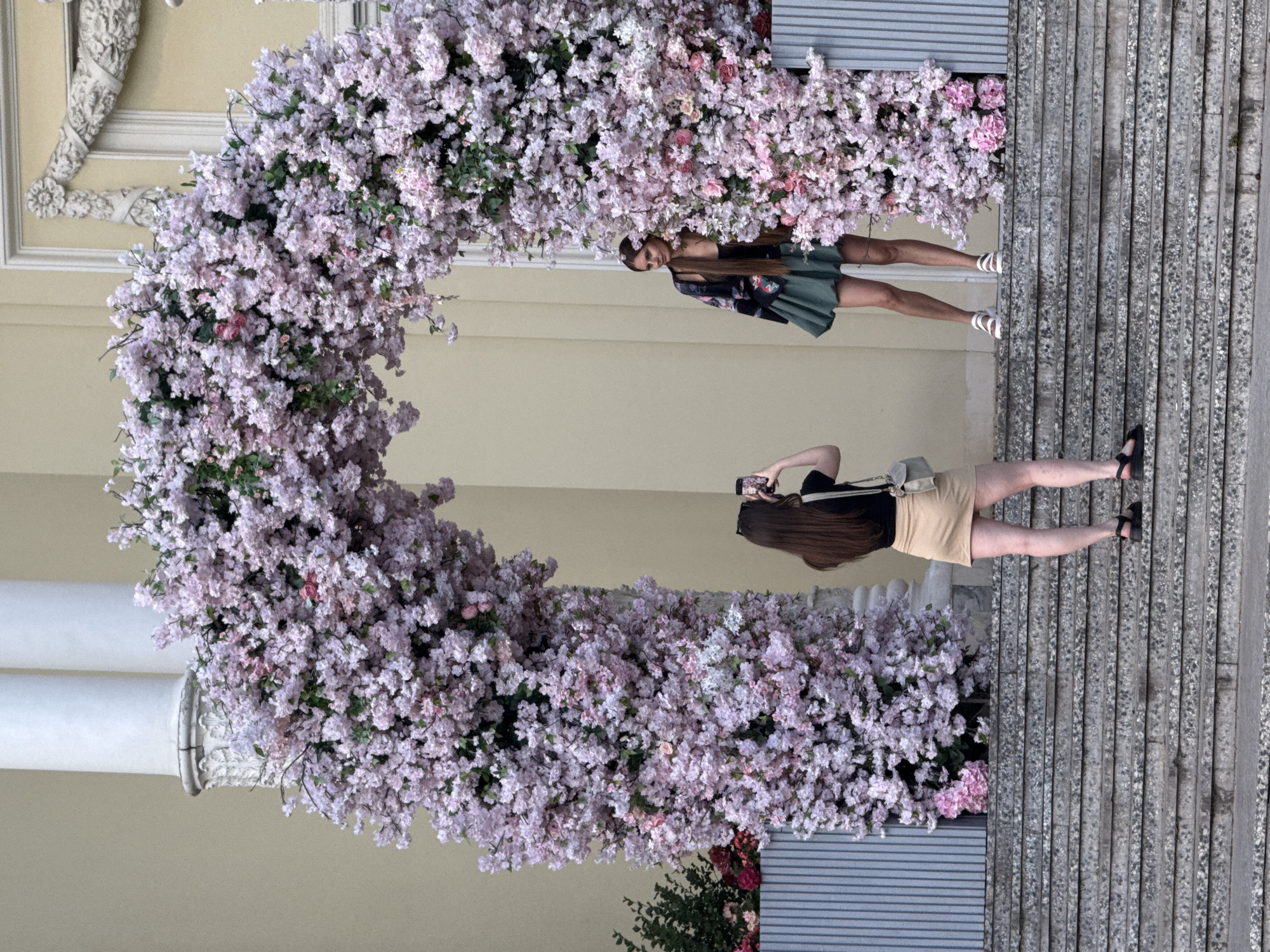 A woman is taking a photograph of another woman standing within a large floral archway. The archway is made of pink and white flowers, with green foliage. The woman being photographed is wearing a black top, a green skirt, and white sandals. She is standing on a set of stone steps. The woman taking the photo is wearing a black t-shirt, a beige skirt, and black sandals. She is also on the stone steps, facing away from the camera and holding up a smartphone. The background consists of a building with a light yellow facade, and two large white columns. The scene appears to be outdoors, possibly in a park or garden. The location is identified as Moscow, Russia.