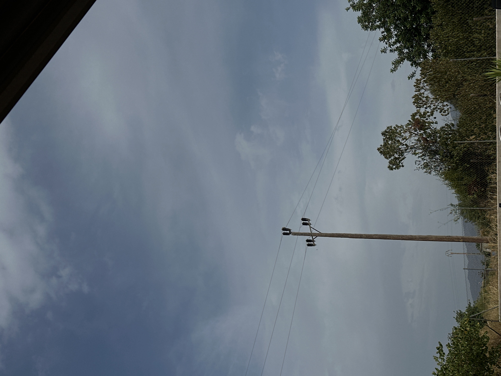 The image depicts an outdoor scene under a cloudy sky. A wooden utility pole with multiple insulators and wires stretches vertically through the center of the frame. To the left of the pole, three parallel wires extend horizontally across the sky. In the foreground, a chain-link fence runs across the bottom of the image, partially obscured by vegetation including a bush on the far left and trees and brush on the right. In the background, a mountainous landscape is visible behind the fence. The location is the city of Santa Maria del Camí, Spain. The scene is devoid of any people or animals.