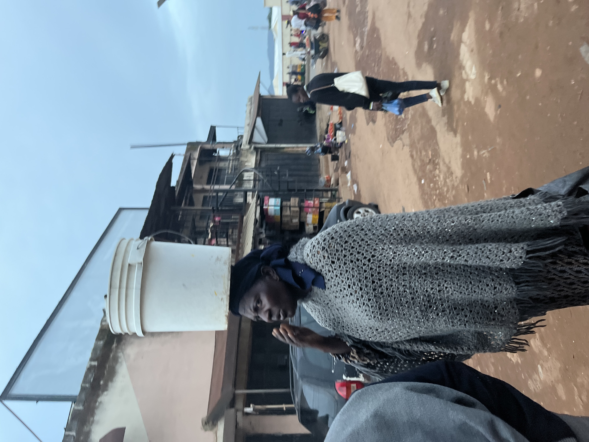 In Jos, Nigeria, a woman carries a white bucket on her head, her right hand near her face. She wears a dark headscarf and a grey crocheted shawl adorned with sequins and fringe. A person in a grey garment stands to her left, partially obscuring her. Behind them, a dark car is parked, and a multi-story building with an outdoor staircase is visible. Further in the background, other people are seen walking in a dusty, reddish-brown urban environment with some market stalls and motorcycles. The sky is overcast.