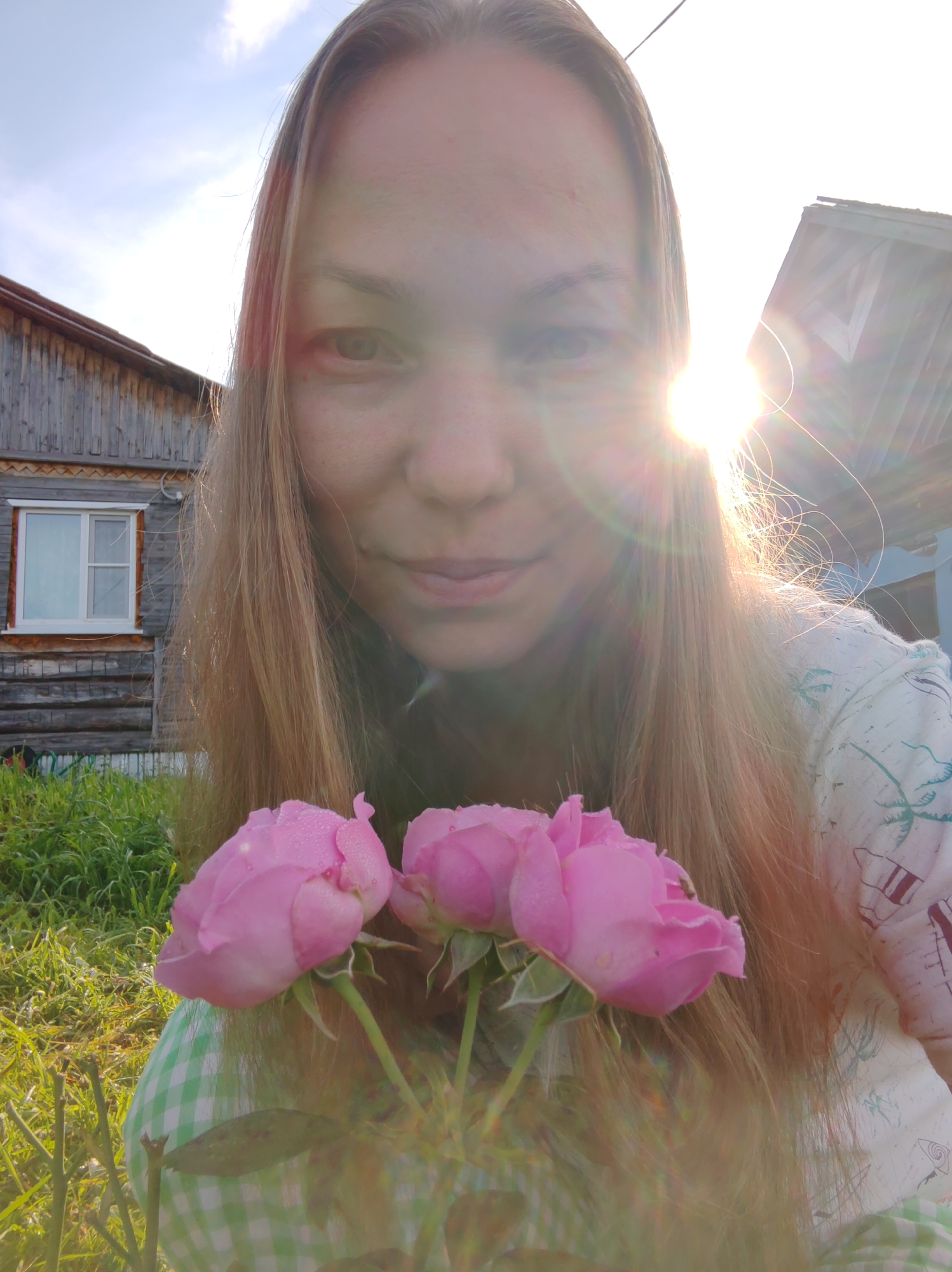 In an outdoor setting, a woman with long blonde hair is taking a selfie. She is holding a small bouquet of three pink roses, which are covered in dew drops. The woman is wearing a light-colored shirt with a pattern of palm trees. Behind her, a weathered wooden house with a white-framed window is visible. The sun is shining brightly, creating a lens flare that obscures parts of the image. The location is identified as Chukhlem, Russia. The overall scene depicts a sunny day with natural elements of a house and garden.