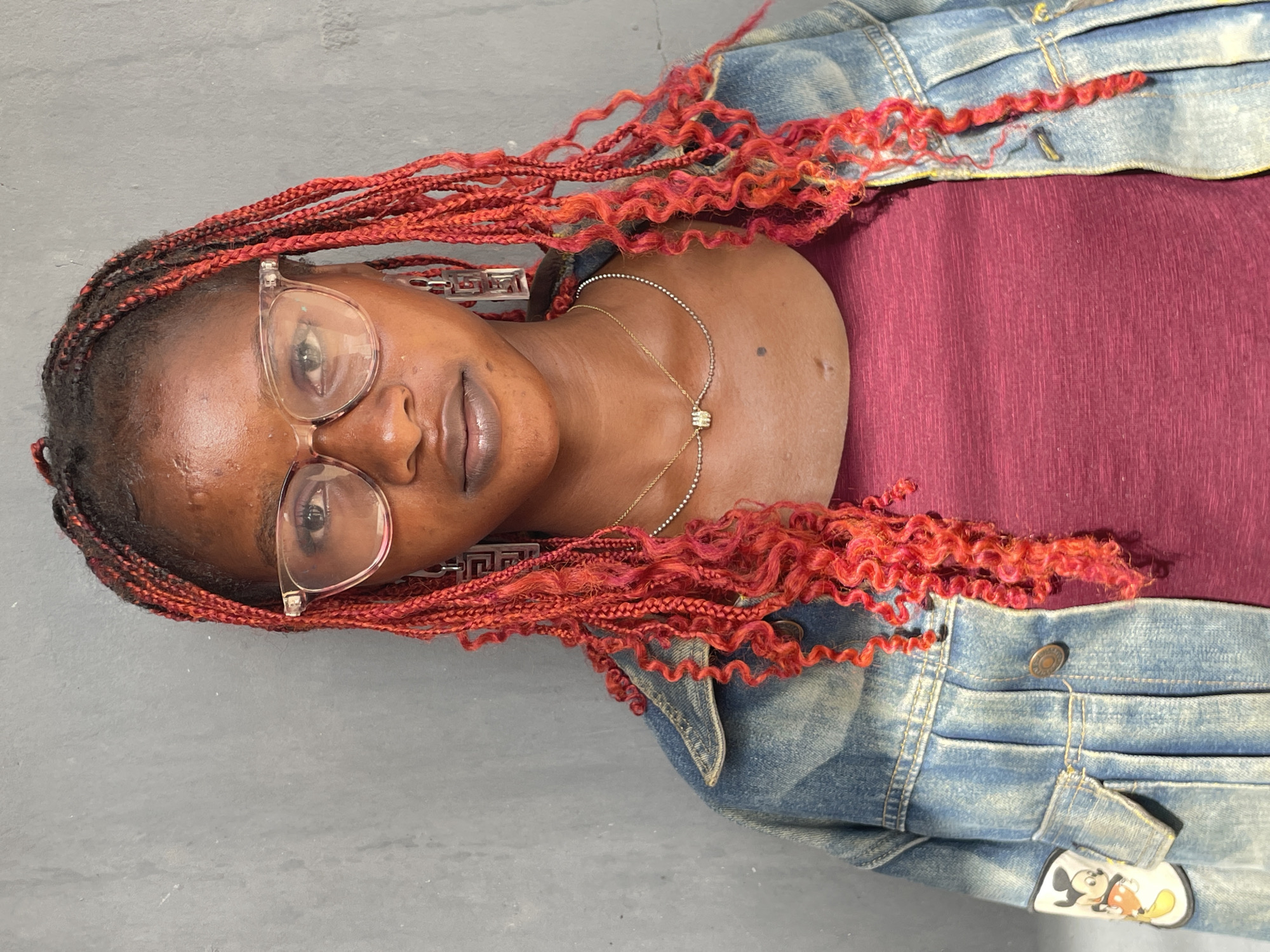 This portrait, rotated 90 degrees clockwise, features a young Black woman positioned against a plain, light gray background. She looks directly at the camera with a calm expression and a slight smile.
Her hair is styled in vibrant red braids, with a mix of textures; some braids are straight, while others are tightly coiled into distinctive curls. She wears clear-framed eyeglasses and prominent geometric earrings, possibly depicting a Greek key pattern. A delicate, gold-toned necklace with a small, square pendant is visible around her neck.
She is dressed in a burgundy or maroon top, partially covered by a light-wash blue denim jacket. A notable detail on the jacket's right sleeve is a small, white patch featuring the animated character Mickey Mouse. The lighting is soft and even, suggesting an indoor setting or shaded outdoor area. The overall mood conveyed is relaxed and self-assured. Based on the provided context, the image was captured in Badagry, Nigeria.