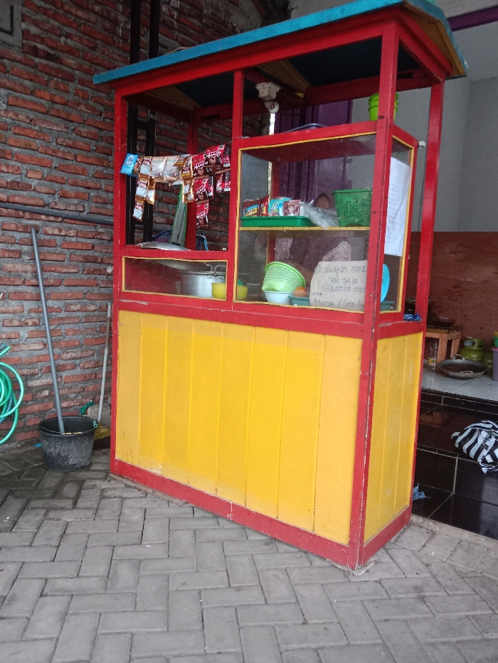 A wooden food stand, painted red and yellow, with a glass front and shelves, is located in front of a brick wall. The stand is in front of a tiled patio area. The stand is empty and there are no people present. There is a black bucket on the ground next to a green garden hose, as well as a broom and a sign on the stand, with writing in Indonesian. The food stand is in the city of Kecamatan Brangsong, Indonesia, at coordinates -6.945 latitude and 110.221 longitude.