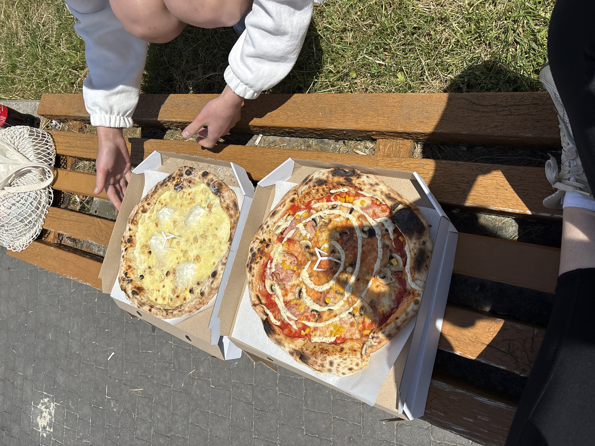 Two pizzas are on a wooden bench in Ternopil, Ukraine. The bench is in a park or outdoor seating area, with cobblestone pavement to the left and grass to the right. Sunlight casts shadows on the bench and the ground.
The pizza in the foreground is in a cardboard box and is topped with tomato sauce, cheese, mushrooms, corn, and a swirl of white sauce. The pizza in the background, also in a box, appears to be a white pizza with cheese and possibly nuts.
Two people are partially visible, one on the right side of the bench reaching towards the pizza. They are wearing casual clothing, including a white sweatshirt and shorts. A white mesh bag and a bottle are also on the bench. The overall scene suggests a casual outdoor meal.