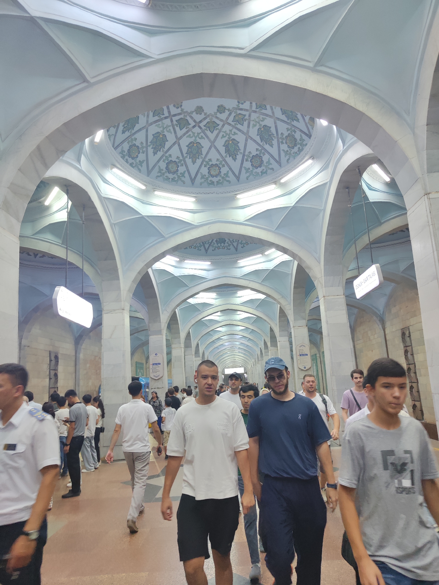 The image depicts the interior of a subway station in Tashkent, Uzbekistan. The architecture features a series of grand arches and a domed ceiling with ornate blue and green floral patterns. Long, fluorescent lights illuminate the cavernous space. A diverse group of people are seen walking in both directions along the platform. In the foreground, several young men are walking towards the viewer. One man in the center foreground wears a blue t-shirt and navy pants, and another on the right wears a grey t-shirt. Other individuals, some in casual wear and one in a white uniform, are also visible, suggesting daily commuter activity. Signs are visible in the background, indicating this is a public transportation hub. The overall impression is of a well-maintained and architecturally significant metro station.