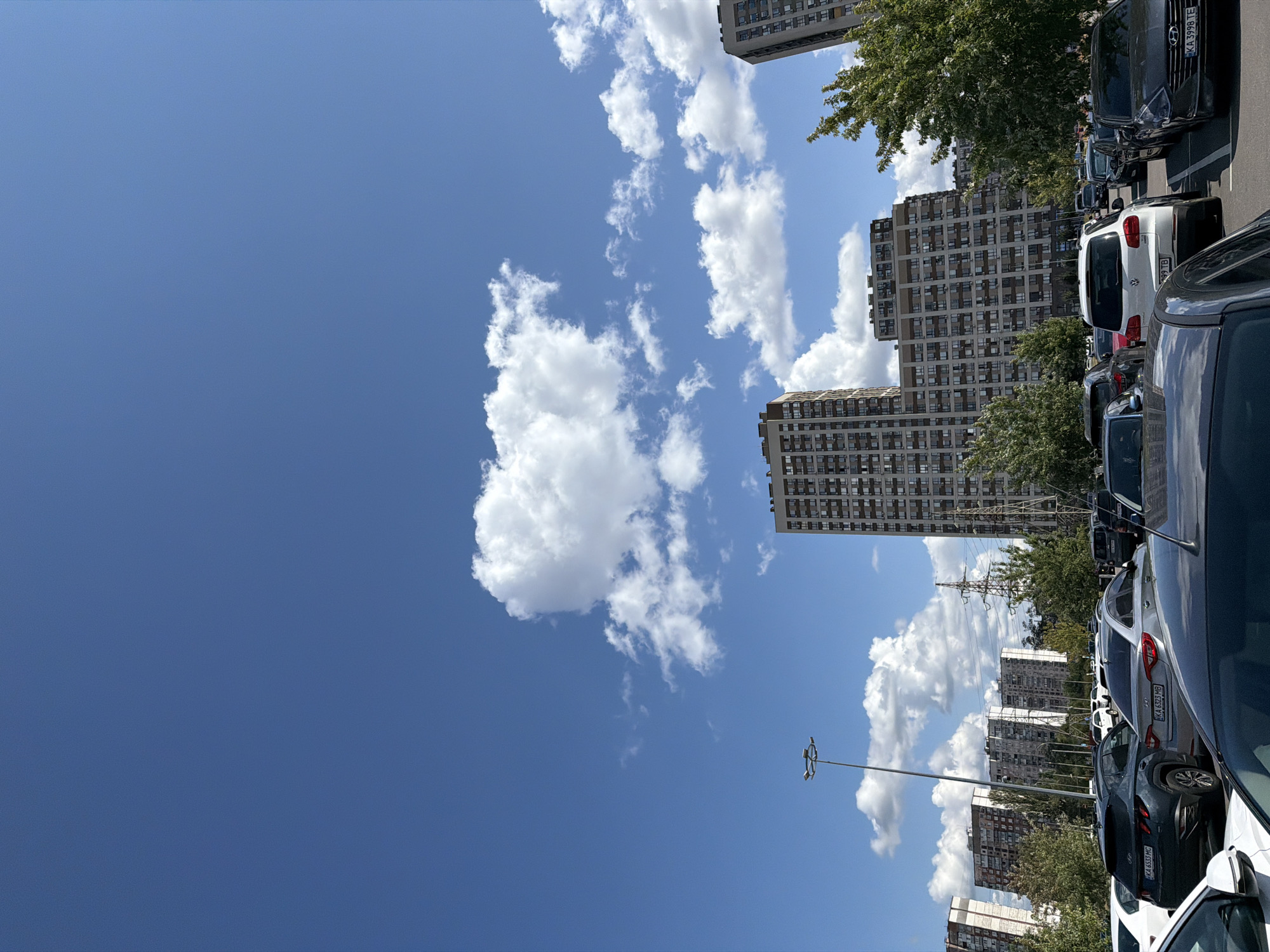 The image captures a wide-angle view of a parking lot filled with numerous cars in a city. The scene is dominated by the bright blue sky with scattered white cumulus clouds. Several modern, multi-story residential buildings are visible in the background, indicating an urban setting. A tall street light stands prominently on the left side of the frame. Power lines and a transmission tower are also present, suggesting utility infrastructure within the city. The foreground is occupied by parked cars, some with visible license plates, hinting at a residential or commercial area in Kyiv, Ukraine. The overall impression is a typical day in a densely populated urban environment.