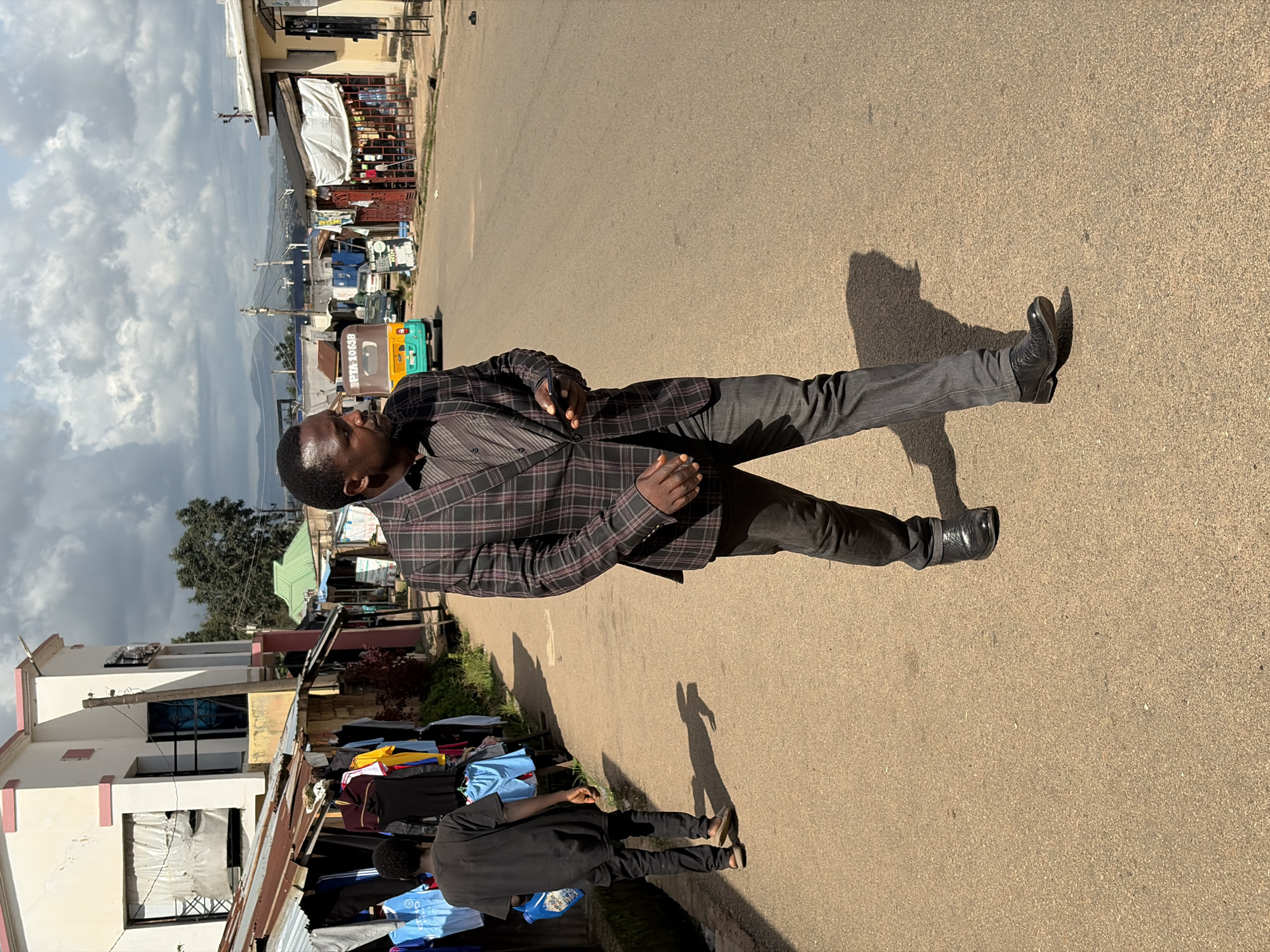 A Black man wearing a plaid blazer, a collared shirt, and dark jeans walks on an asphalt road in Jos, Nigeria. He holds a mobile phone in his right hand. To his left, a younger Black man wearing a black t-shirt and black jeans walks away from the camera, carrying a blue detergent bottle. He is walking past a row of market stalls selling clothing, with various garments displayed on racks. The market stalls are covered with corrugated metal roofs and are adjacent to a multi-story building. In the background, a green-roofed structure is visible, along with other buildings, parked vehicles including a tricycle, and utility poles with power lines. The sky is cloudy.