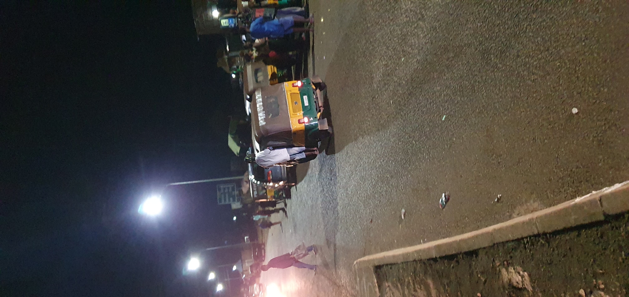 At night in Barakin Angundi, Nigeria, a street scene unfolds under the glow of streetlights. The asphalt road is wet, reflecting the lights and creating a shimmering effect. Several vehicles are present, including a yellow and green tuk-tuk in the foreground with visible writing on its side. A dark-colored van is parked behind it. In the background, more vehicles and various structures line the street, suggesting a commercial or transit area.
People are scattered across the scene. A man wearing a white shirt and blue shorts stands near the tuk-tuk. Further to the left, a person in a light-colored outfit walks across the road, carrying bags. Other individuals are visible in the background, some standing near the vehicles and structures, their actions unclear in the low light. The overall atmosphere is that of a busy street at night, possibly a transportation hub or market area, illuminated by artificial lighting. The presence of debris on the wet road indicates recent activity or potential neglect.