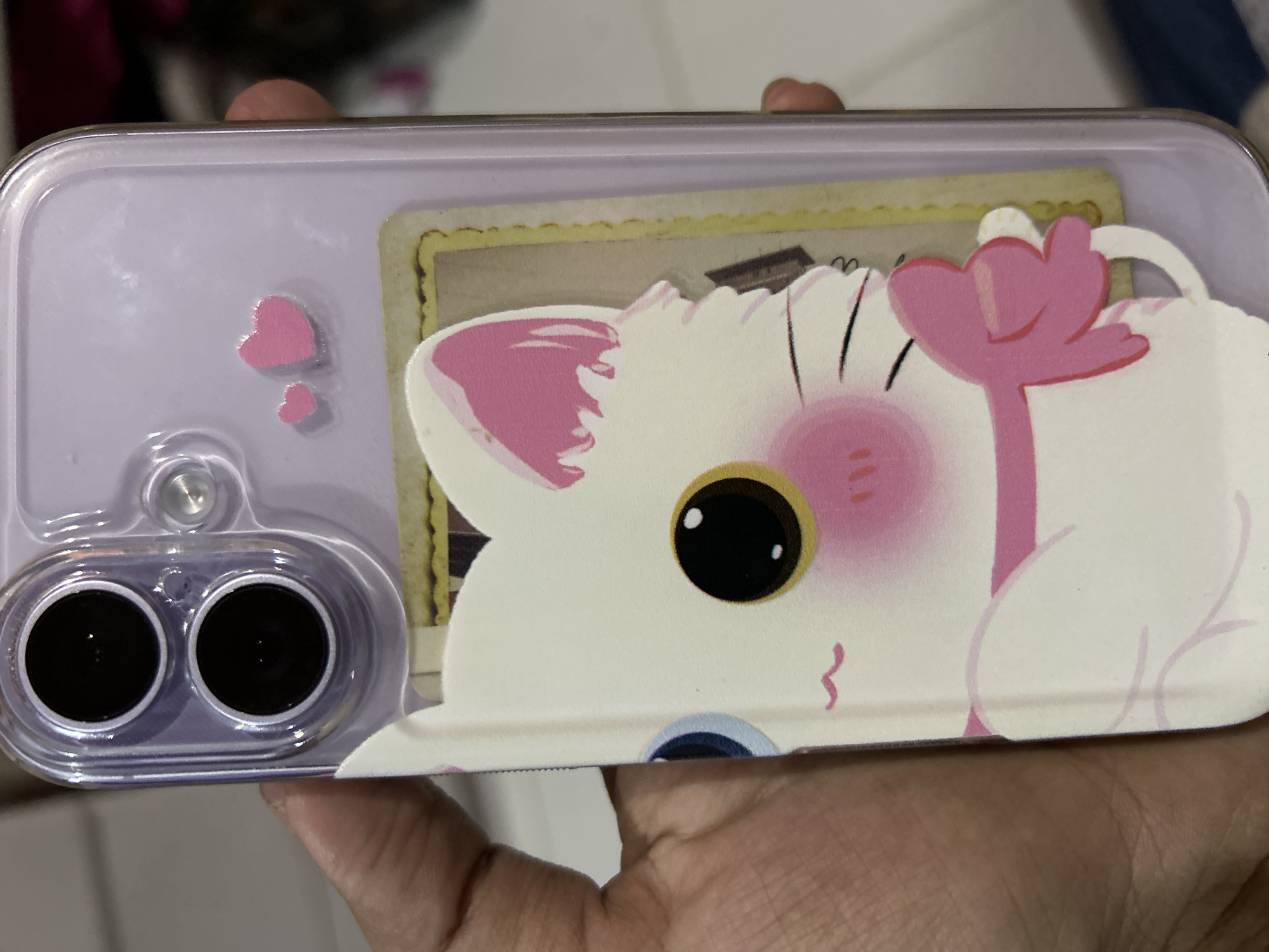 A person's hand holds a clear, purple phone case with a cartoon cat design. The phone case is on a smartphone, and the camera lenses are visible at the top. The cartoon cat design features a white cat with pink accents on its ears and a pink bow around its neck. Two small pink hearts are also adhered to the phone case. A card with a yellow border is visible behind the cat design. The background is blurred and appears to be an indoor setting. The image was taken in Depok, Indonesia.