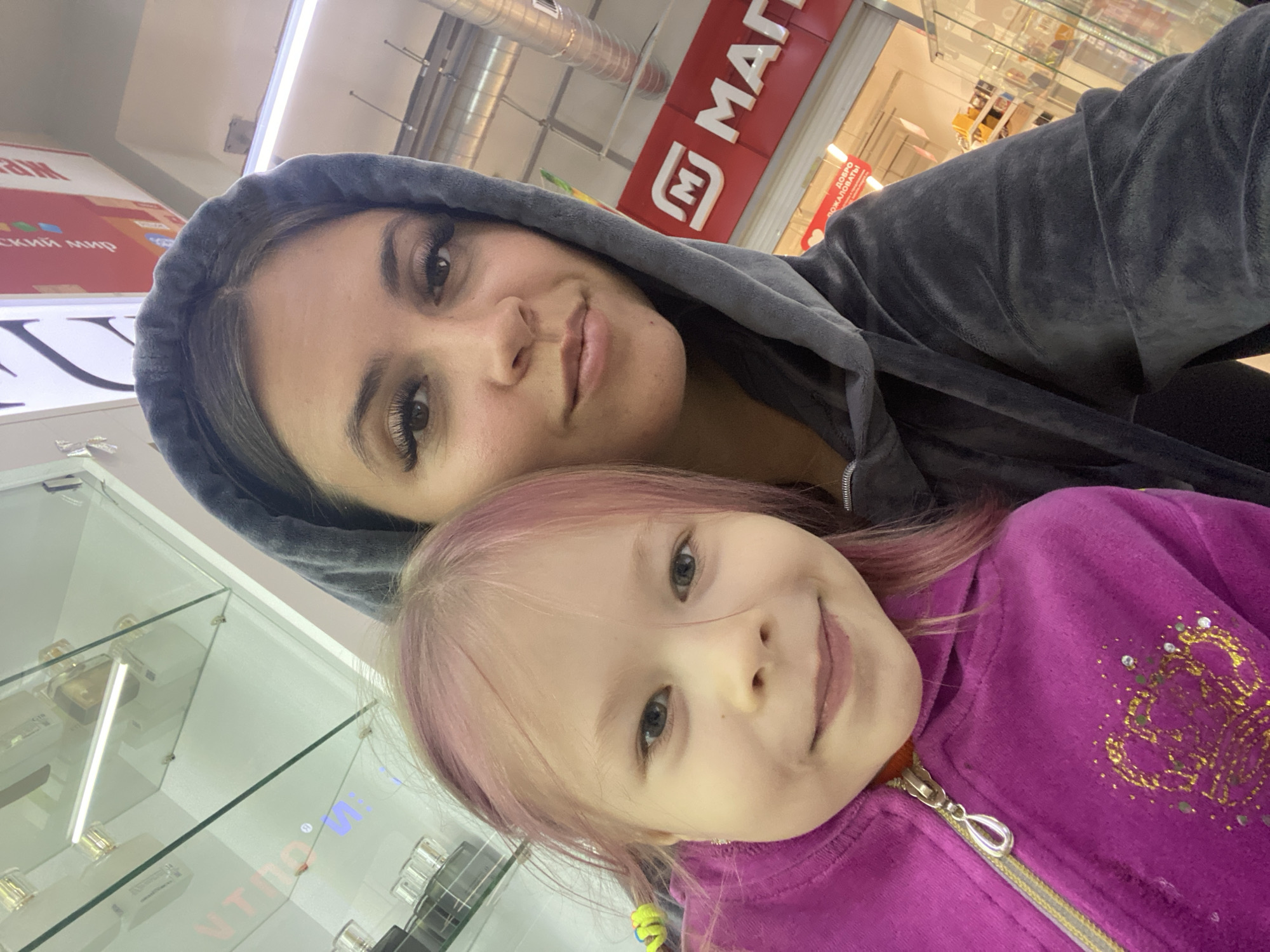 A selfie is taken by two people, a woman and a young girl, in what appears to be a retail environment. The woman, on the right, is wearing a dark gray hooded sweatshirt. She has dark hair, visible makeup including dramatic eyelashes, and is looking directly at the camera. The young girl, on the left, has light pink hair styled in two pigtails with yellow ties. She is wearing a bright magenta hooded sweatshirt with a zipper and a decorative design on the front. She is smiling and looking at the camera. In the background, a red sign with white lettering, possibly a store name, is visible. Above the sign, a light fixture illuminates the space. Shelves with products are also discernible in the background. The scene is indoors, with bright, artificial lighting.
The provided metadata indicates that the location is Chaikovsky, Russia. While the specific store is not identified, the presence of merchandise and signage suggests it is a commercial establishment.