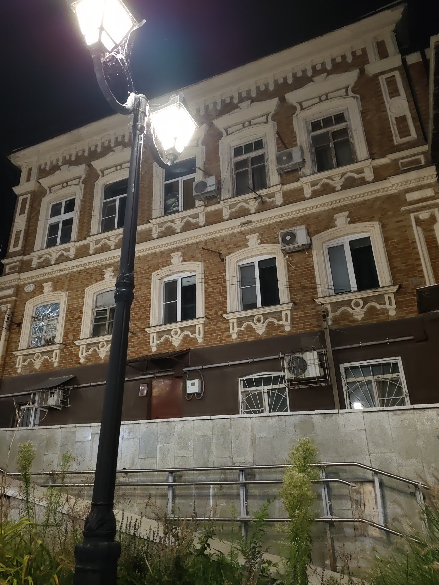 The image shows a lamppost in front of a brick building at night. The lamppost has two lamps that are lit, illuminating the surrounding area. The building appears to be old, with decorative white trim around the windows. There are several windows on the facade of the building, some of which are open. There are also air conditioning units mounted on the walls. In the foreground, there is some vegetation and a metal railing. The scene is dimly lit, with the lamppost being the primary source of light. The city of Rostov-on-Don is known for its historical architecture, and this building could be an example of that. The image captures a typical urban night scene, with a focus on the interplay of light and shadow on the building's facade.
