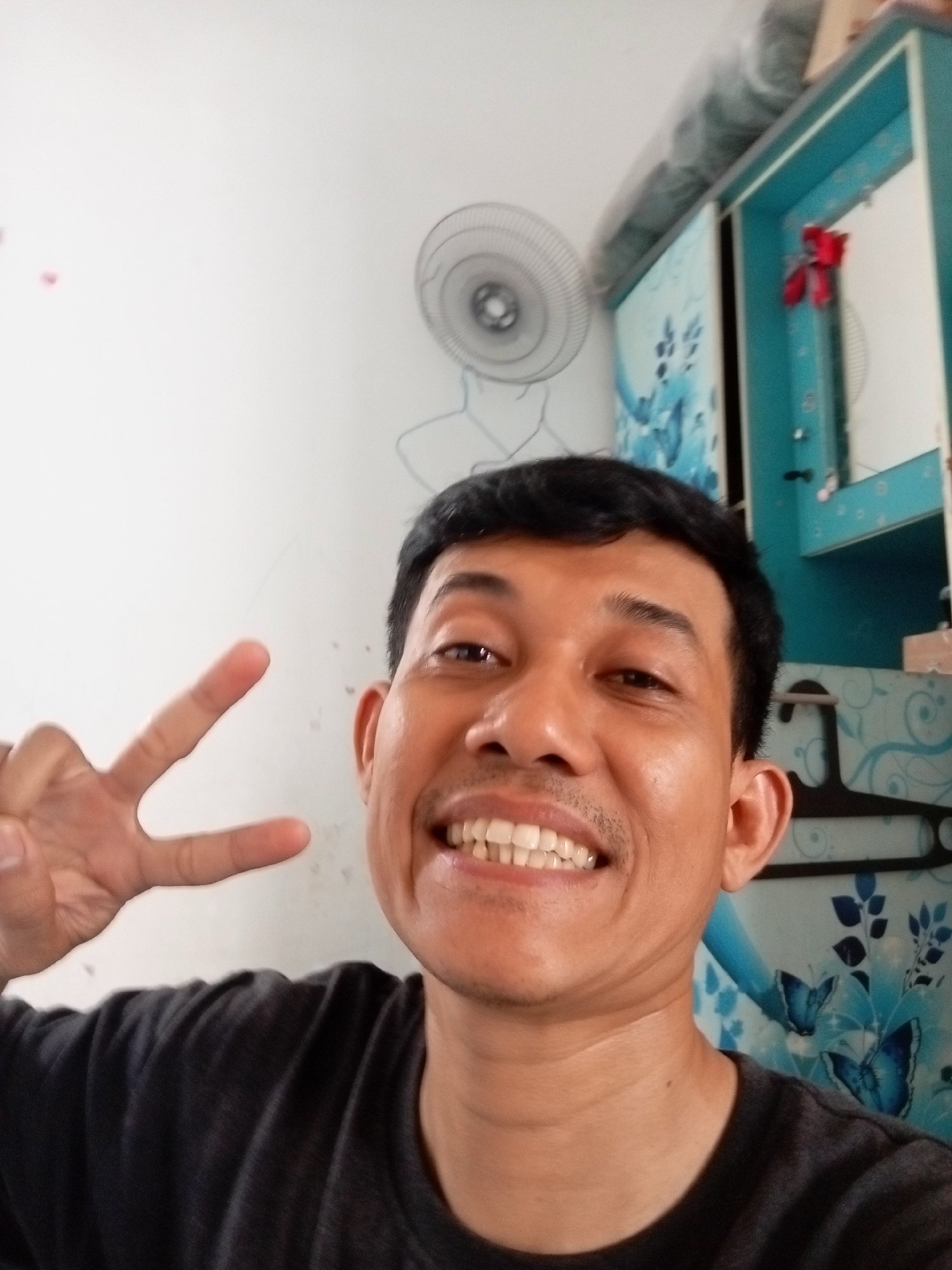 In Batam, Indonesia, a close-up selfie shows a man with dark hair and a light brown complexion smiling and making a peace sign with his left hand. He is wearing a dark gray t-shirt. Behind him, a white wall is visible, with a round electric fan mounted on it. To the right, a blue cabinet with floral and butterfly motifs is present, featuring a red ribbon decoration on its upper section.