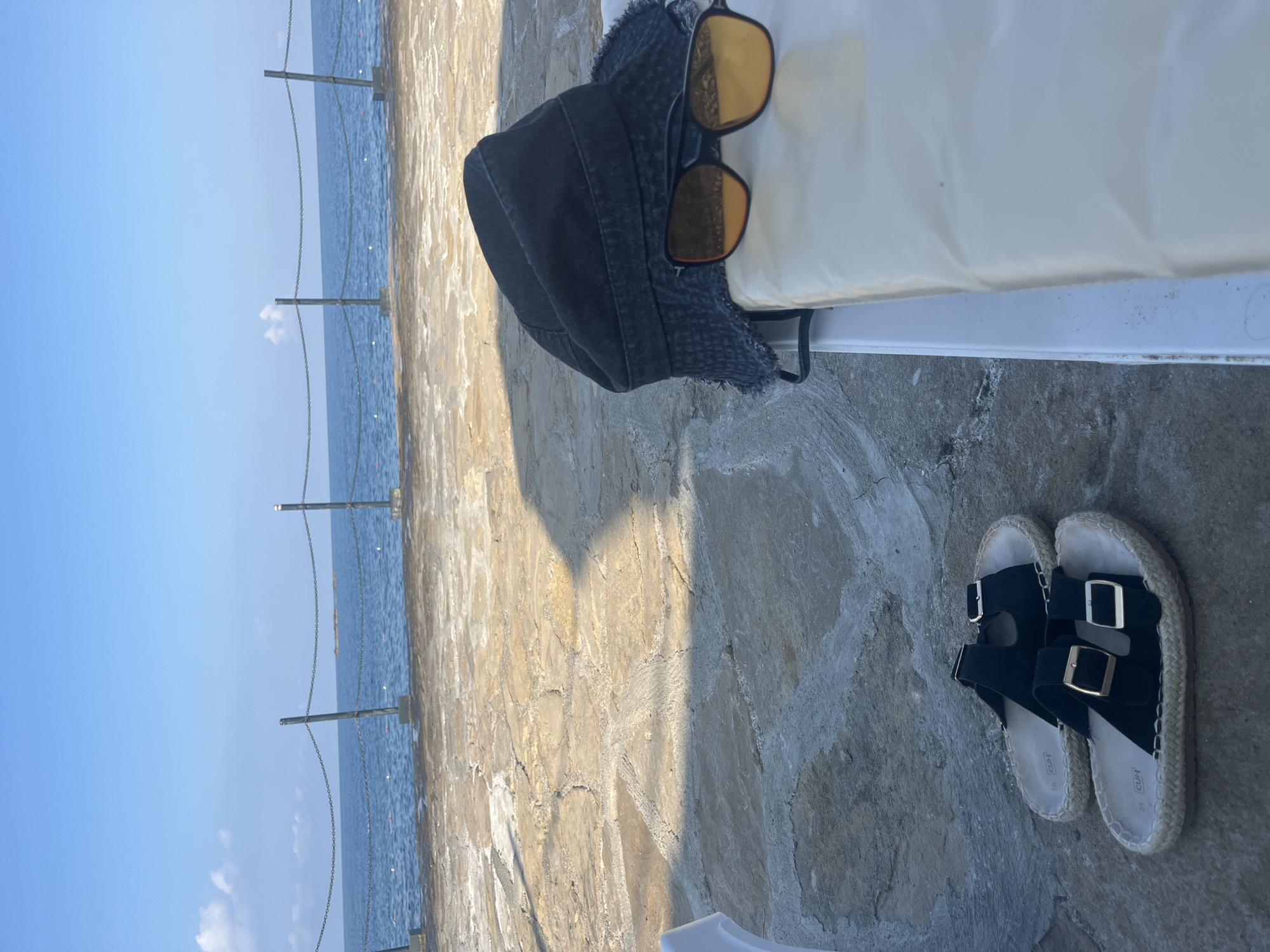 This is a close-up, eye-level shot of beach accessories on a stone patio next to the sea. The scene is oriented vertically, so the horizon line appears on the left side of the frame. The subjects are a pair of black sandals with gold buckles and a black denim bucket hat with yellow-tinted sunglasses resting on top. The sandals are arranged side-by-side, with the left sandal slightly overlapping the right. The hat is positioned behind the sandals, with its brim facing towards the top of the frame. Behind the hat and sunglasses is a white object, possibly a beach chair or towel, which is mostly out of focus.
The background on the left consists of the calm, blue sea under a clear, bright sky. A chain-link fence with metal posts runs parallel to the shore, separating the patio from the water. The stone patio itself, on the right, is made of large, irregularly shaped rocks with visible cracks and grout lines. A shadow is cast across the patio from an unseen object, suggesting it is daytime with the sun positioned to the right and slightly behind the viewer. The overall mood is relaxed and suggests a holiday or leisure activity. The location is identified as Alanya, Turkey. No visible text is present in the image.