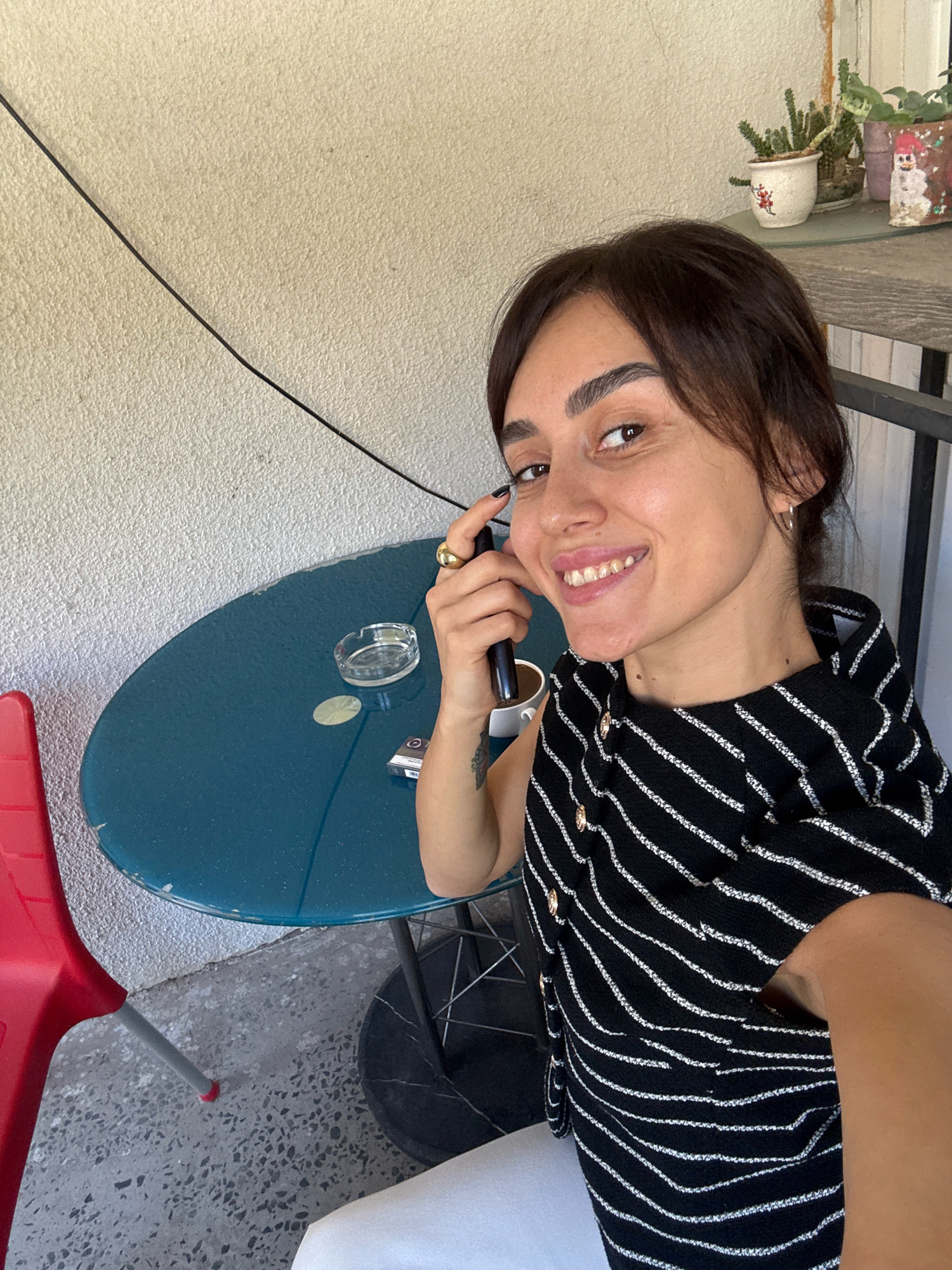 A woman is taking a selfie at an outdoor table with a blue surface. She is smiling and holding a dark-colored electronic device to her ear. She is wearing a black and white striped top with gold buttons. A glass ashtray, a pack of cigarettes, and a white cup are on the table. A red chair is to the left of the table, and a black metal chair is visible below the table. A wall with a rough texture is behind the woman and table. In the background, on a shelf, are several potted plants and a decorative snowman. The scene appears to be on a balcony or patio. The image was taken in Tbilisi, Georgia.