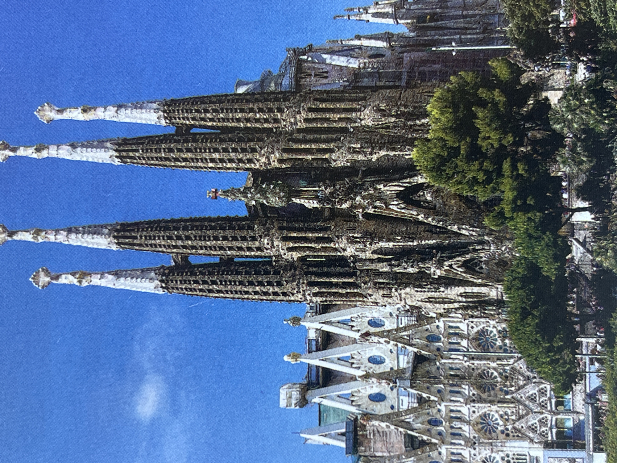 The image shows the Sagrada Familia basilica in Barcelona, Spain, under a bright blue sky. The basilica, a UNESCO World Heritage Site, is characterized by its intricate and unique architectural style, with towering spires adorned with symbolic sculptures. Several spires are visible in the foreground, featuring detailed stonework and Gothic-inspired elements. The façade of the basilica exhibits elaborate carvings and pointed arches, with a series of ornate windows and decorative features. To the left of the main structure, a section of the basilica with triangular gables and circular windows is visible. In the lower part of the image, lush green trees and foliage frame the base of the basilica, and a crowd of people can be seen in the distance, suggesting it is a popular tourist destination. The overall scene conveys a sense of grandeur and architectural magnificence against a clear sky.