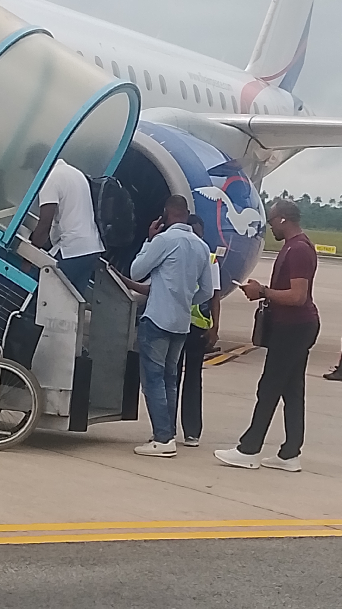In Orerokpe, Nigeria, at an airport tarmac, several individuals are observed boarding an airplane. A person in a white shirt is ascending a mobile staircase, followed by another person in a light blue shirt and jeans, who is speaking on a mobile phone. Behind them, a person wearing a high-visibility yellow vest is also visible. To the right, a man in a maroon shirt and black pants is holding a mobile phone and looking down at it while wearing earbuds. The aircraft, with a distinctive blue and white logo on its tail, is parked on the tarmac. The ground is paved, with yellow lines indicating a boundary. The overall scene depicts the process of passengers boarding a commercial flight.