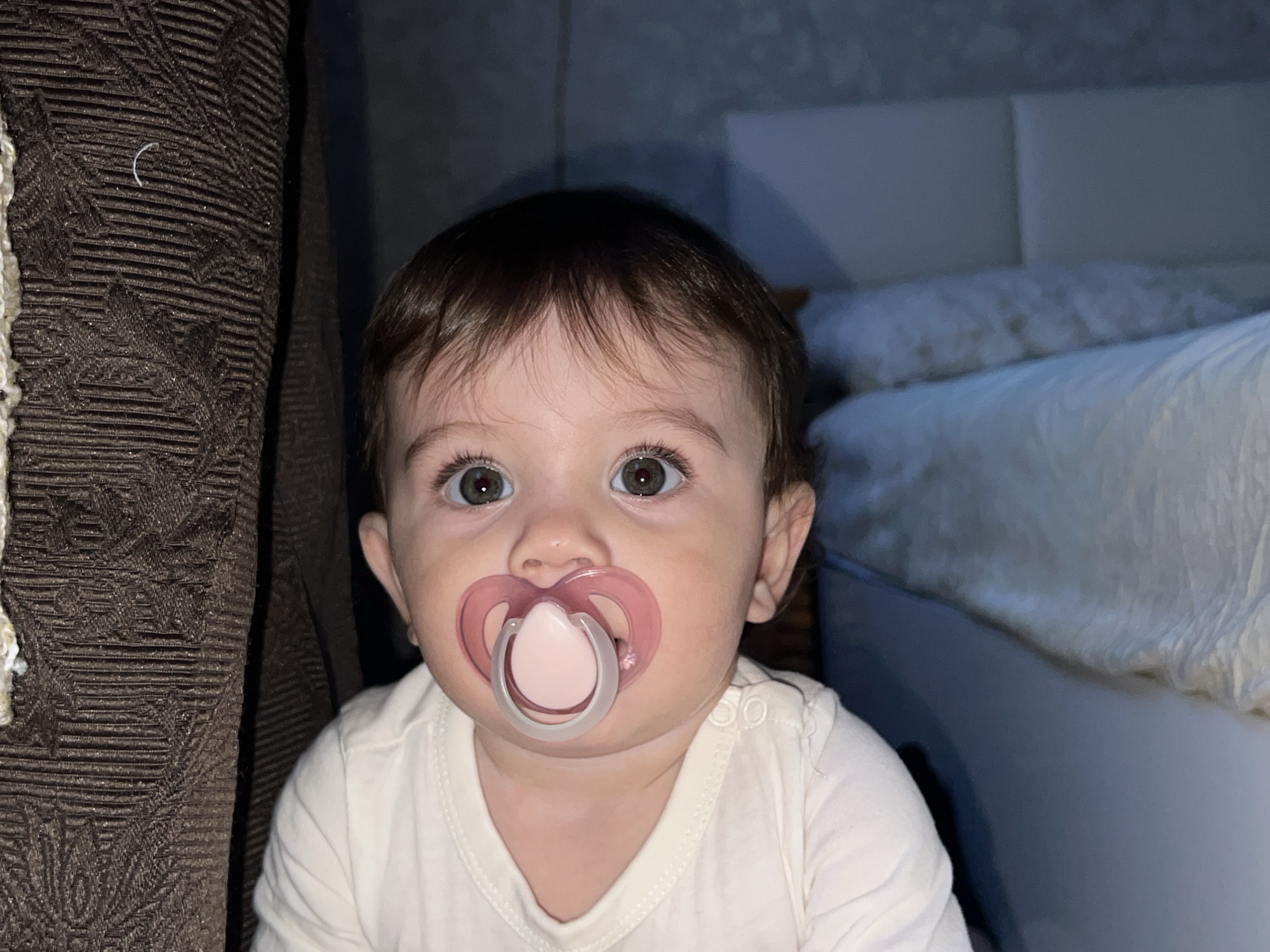 An infant or toddler, with dark brown hair and light green or hazel eyes, is depicted in a close-up, head-on view. The individual is wearing a light cream-colored short-sleeved onesie or t-shirt with snap closures visible on the right shoulder. A translucent pink pacifier with a lighter pink bulbous nipple is inserted into the individual's mouth. The individual is looking directly forward towards the camera. To the left, a dark brown patterned fabric, possibly a curtain or upholstery, is visible, with a small white curved fiber resting on its surface. To the right and behind the individual, a padded white or light gray headboard and white bedding, including pillows and a duvet, are present, indicating an indoor bedroom setting. The specific location in the city of Elena, Bulgaria, is not discernible from the visual evidence. No other individuals or interactions are visible.