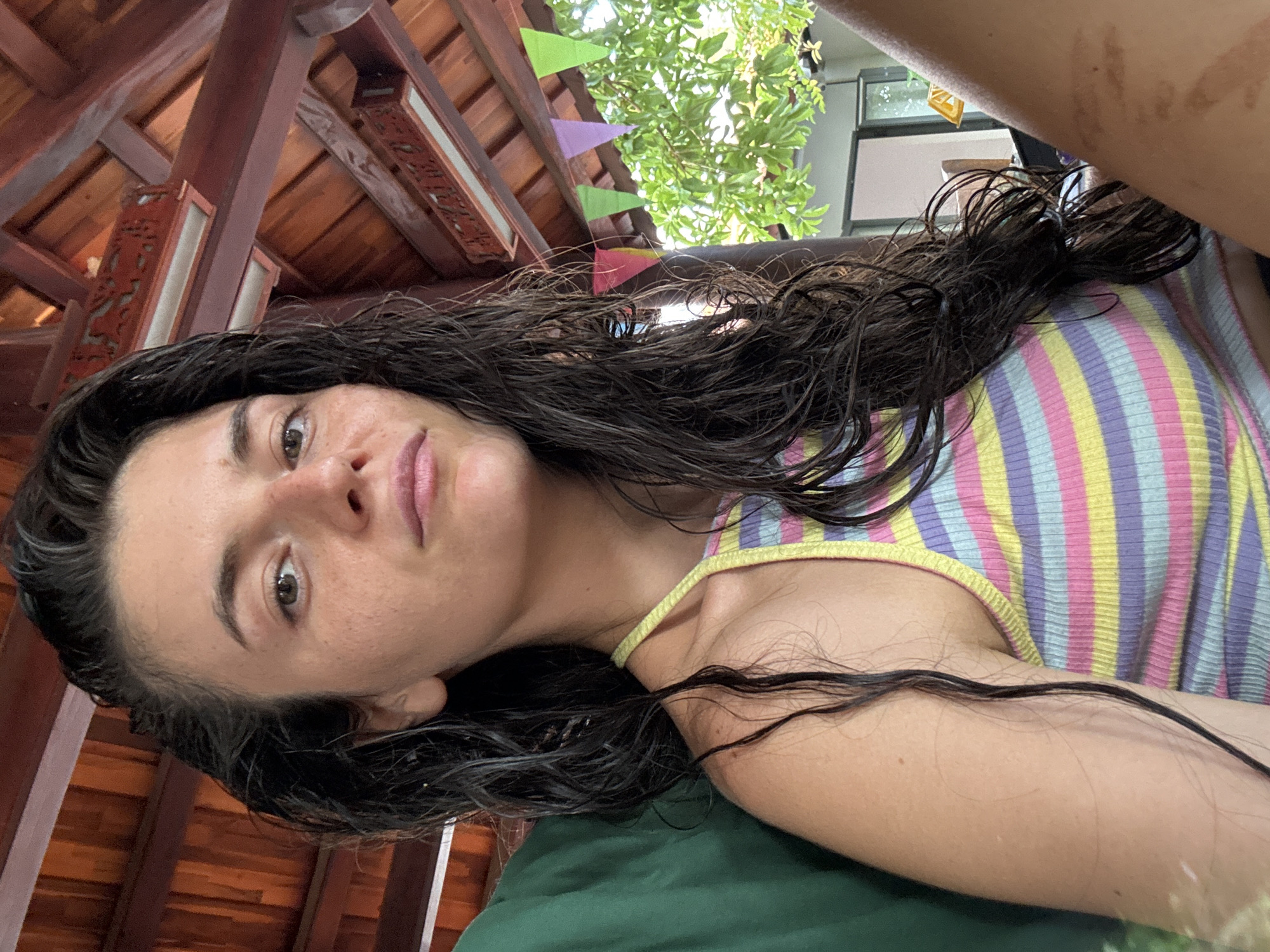 A young woman with long, dark, wet hair and freckles on her face poses for a selfie indoors. She is wearing a colorful striped tank top. The background shows a wooden ceiling with decorative elements and a string of triangular flags. Green foliage and a glimpse of an outdoor setting are visible to the right. The setting appears to be a covered porch or pavilion, typical of the beach resorts found in Mũi Né, Vietnam.