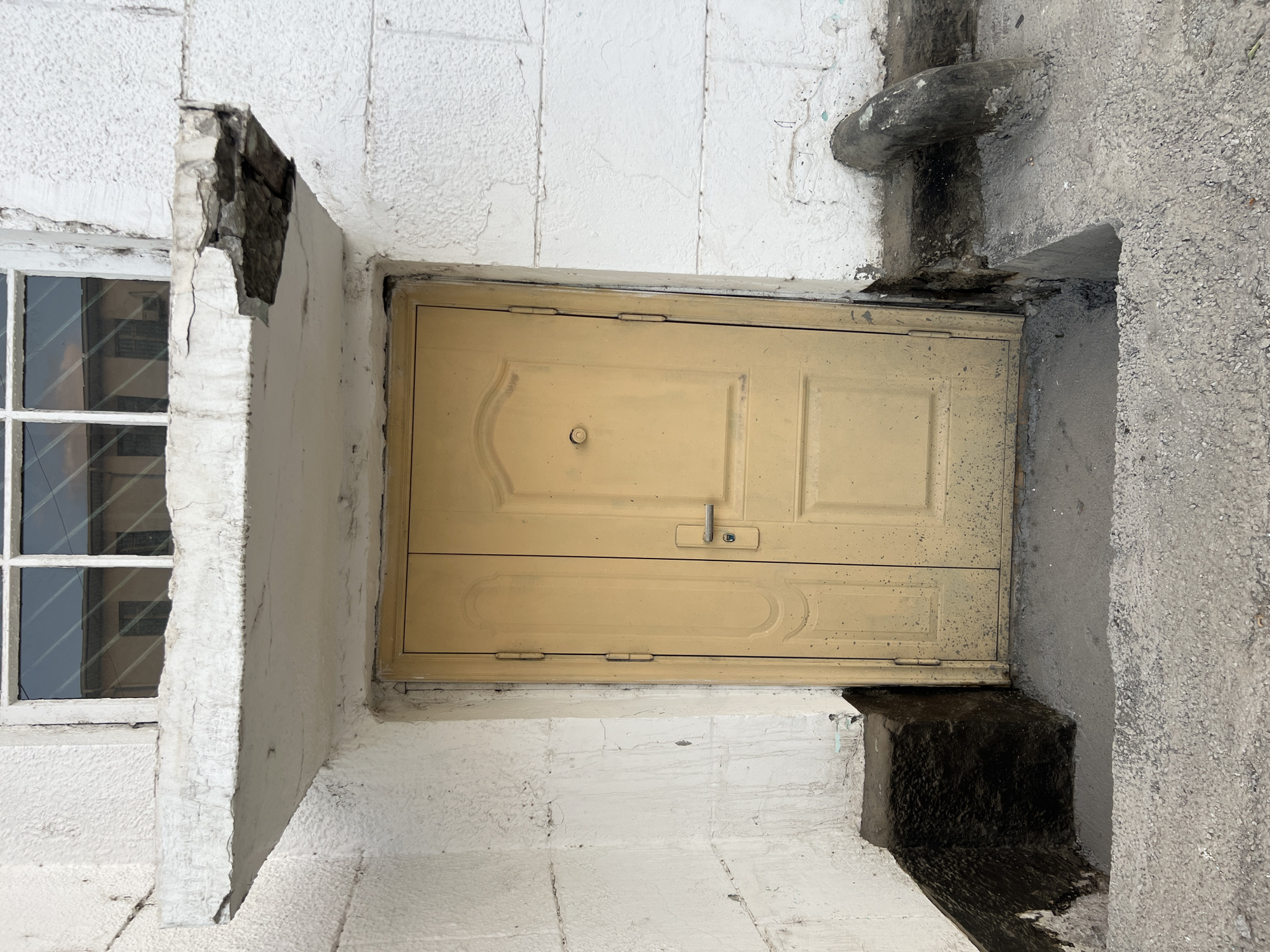 The image shows a dilapidated entrance to a building. A yellow double door, weathered and scratched, is centrally located. Above the door, a concrete overhang shows significant damage with a large piece missing from the right side. A window with a grid is visible above the overhang. The building's exterior walls are white and appear to be made of rough, textured blocks. The ground in front of the door is a mix of concrete and gravel, with some dark, possibly tar-coated, raised areas flanking the entrance. A curved, dark pipe protrudes from the wall to the right of the door. The scene is likely in Osh City, Kyrgyzstan, given the context.