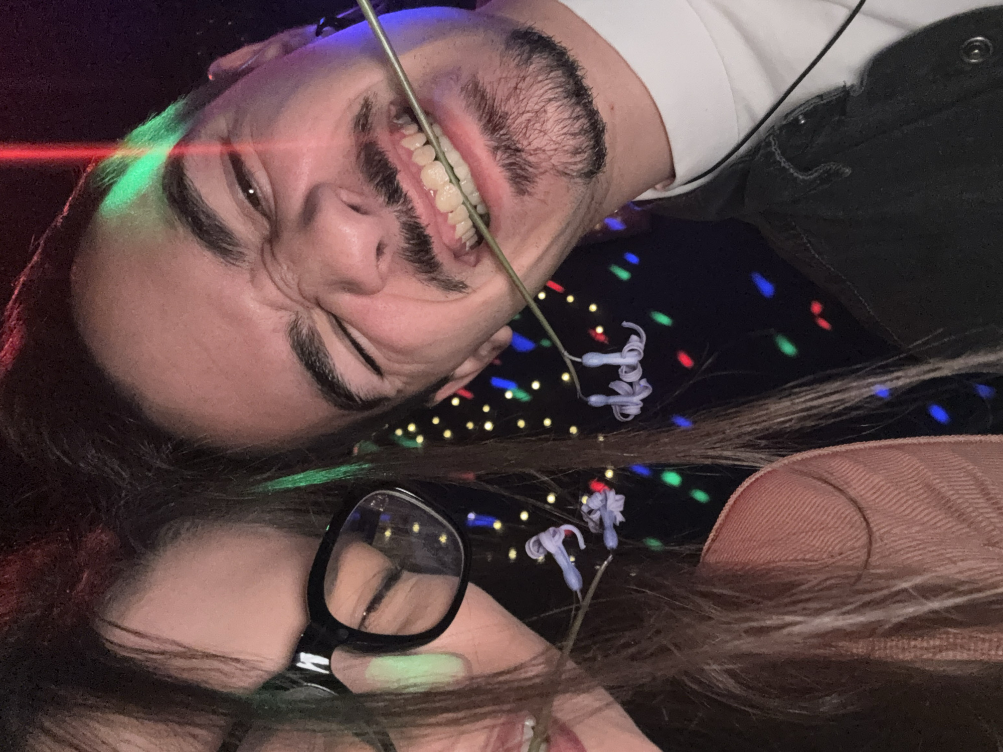This close-up image depicts two individuals, a male and a female, in a dimly lit indoor environment in Sofia, Bulgaria. The male, positioned on the right, has dark hair, a beard, and a mustache, displaying an exaggerated grin with visible teeth and scrunched facial features. Red and green light beams are visible across his face and hair. He wears a white collared shirt beneath a dark-colored jacket. The female, partially visible on the left, wears black-framed glasses and has long, dark hair with subtle green streaks. A thin, green, flexible stem or wire is held in the mouths of both individuals, creating a connection between them. Three small, white, decorative, flower-like objects are attached to this wire near the center. The background is dark and features numerous out-of-focus, multi-colored lights (red, green, blue, yellow), consistent with club or event lighting.