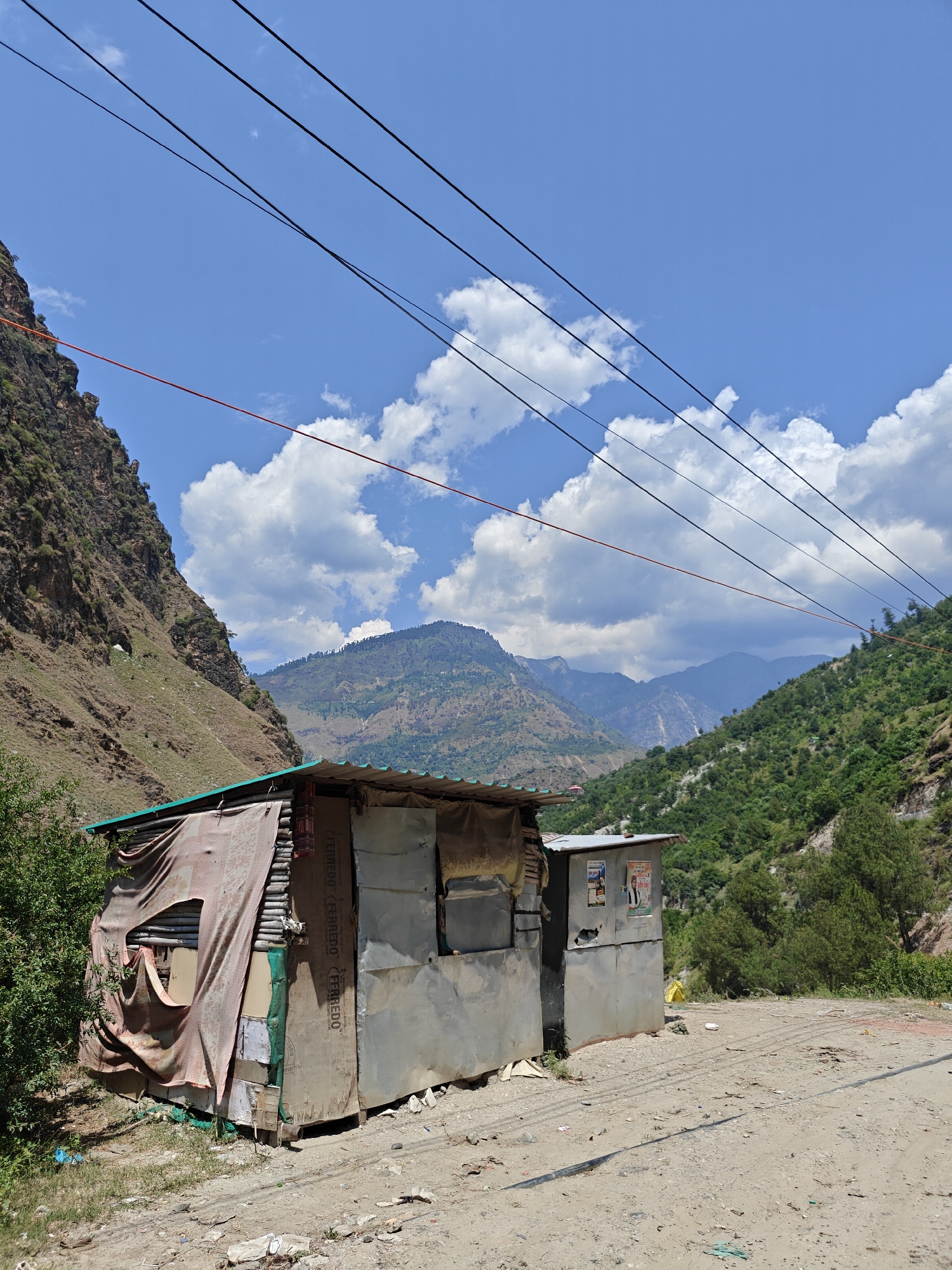 The image displays a mountainous landscape in Shat, India, under a bright, clear blue sky dotted with scattered white cumulus clouds. A dirt road runs through the foreground and extends into the distance.
On the right side of the road are two makeshift structures. The larger one on the left is constructed from various materials, including corrugated metal for the roof and what appears to be repurposed wooden boards or plywood, bearing the repeated text "FERREDO." A patterned, reddish-brown fabric or blanket is draped over the front and side, covering a small opening. Adjacent to this, a smaller, simpler structure is visible, primarily built from corrugated metal. Several posters are affixed to its side, featuring images of men; one prominent poster shows a man wearing a white cap, likely a political figure or advertisement.
The setting is a valley or gorge, with steep, rugged mountainsides rising on either side. The mountain on the immediate left is predominantly rocky and barren, while the mountains in the mid-ground and distant background are covered with dense green vegetation and trees. Overhead, multiple black power lines and a single red line traverse the sky, implying infrastructure connecting the remote area.
The scene suggests a midday or early afternoon setting due to the bright, direct sunlight. No people or active events are depicted, presenting a static and tranquil, yet perhaps isolated, environment. Small pieces of litter are visible on the dirt ground near the structures.
