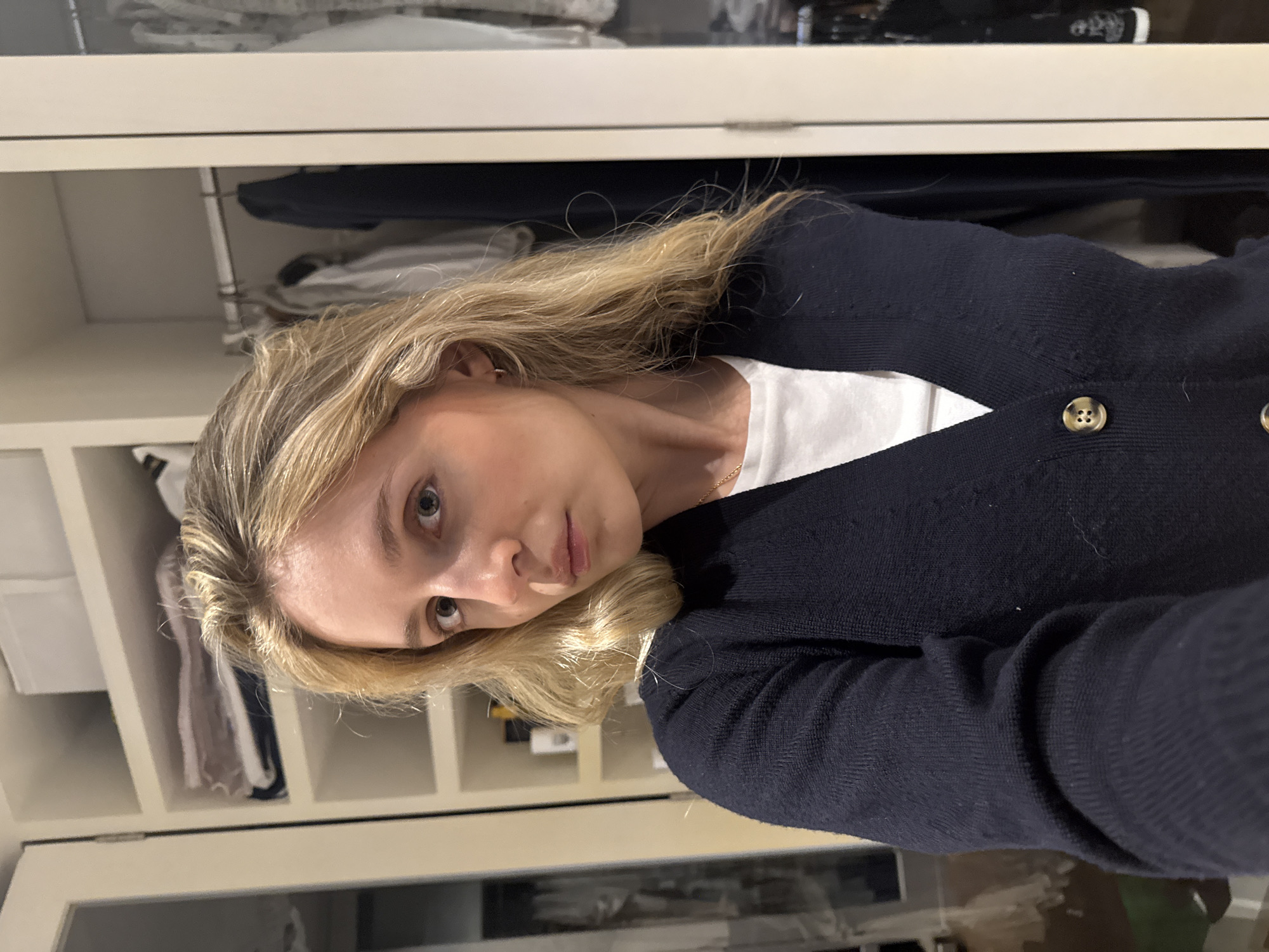 A young Caucasian woman with blonde hair is taking a selfie in a closet. She is wearing a navy blue cardigan over a white t-shirt, and a thin gold necklace. The closet behind her is organized with shelves of folded clothes on the left and hanging garments on the right. The woman has a neutral expression and is looking directly at the camera. The environment appears to be a well-lit indoor space, possibly a bedroom or dressing area. There is no visible indication of the location being in Mexico City.