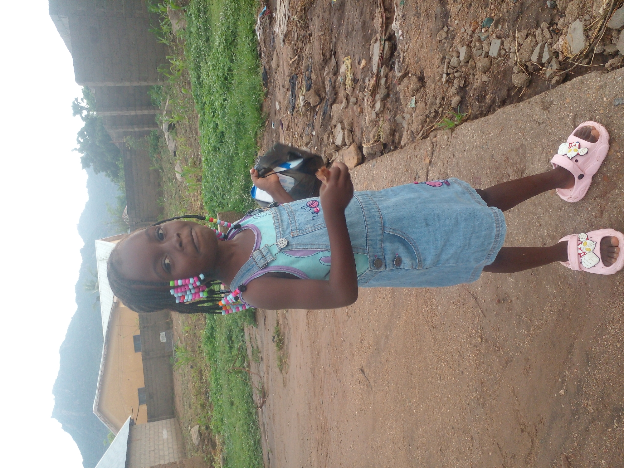 A young Black girl stands on a dirt path in front of a concrete curb. She is wearing a light blue denim overall dress with a light blue tank top underneath. The dress has pockets on the front with a pink embroidered bee on the left pocket. She has braids in her hair decorated with colorful beads. She is holding a black plastic bag in her right hand and a small brown object in her left hand. She is wearing pink sandals with white flower designs on them. In the background, there are several buildings, some with yellow walls and corrugated metal roofs. Behind the buildings, a large mountain is visible under a hazy sky. The ground around the path is covered in dirt, rocks, and some patchy green vegetation. There is also debris scattered on the ground to the right of the girl. The overall scene suggests an outdoor environment, possibly in a rural or semi-urban area of Langtang, Nigeria, given the architectural style of the buildings and the natural landscape.