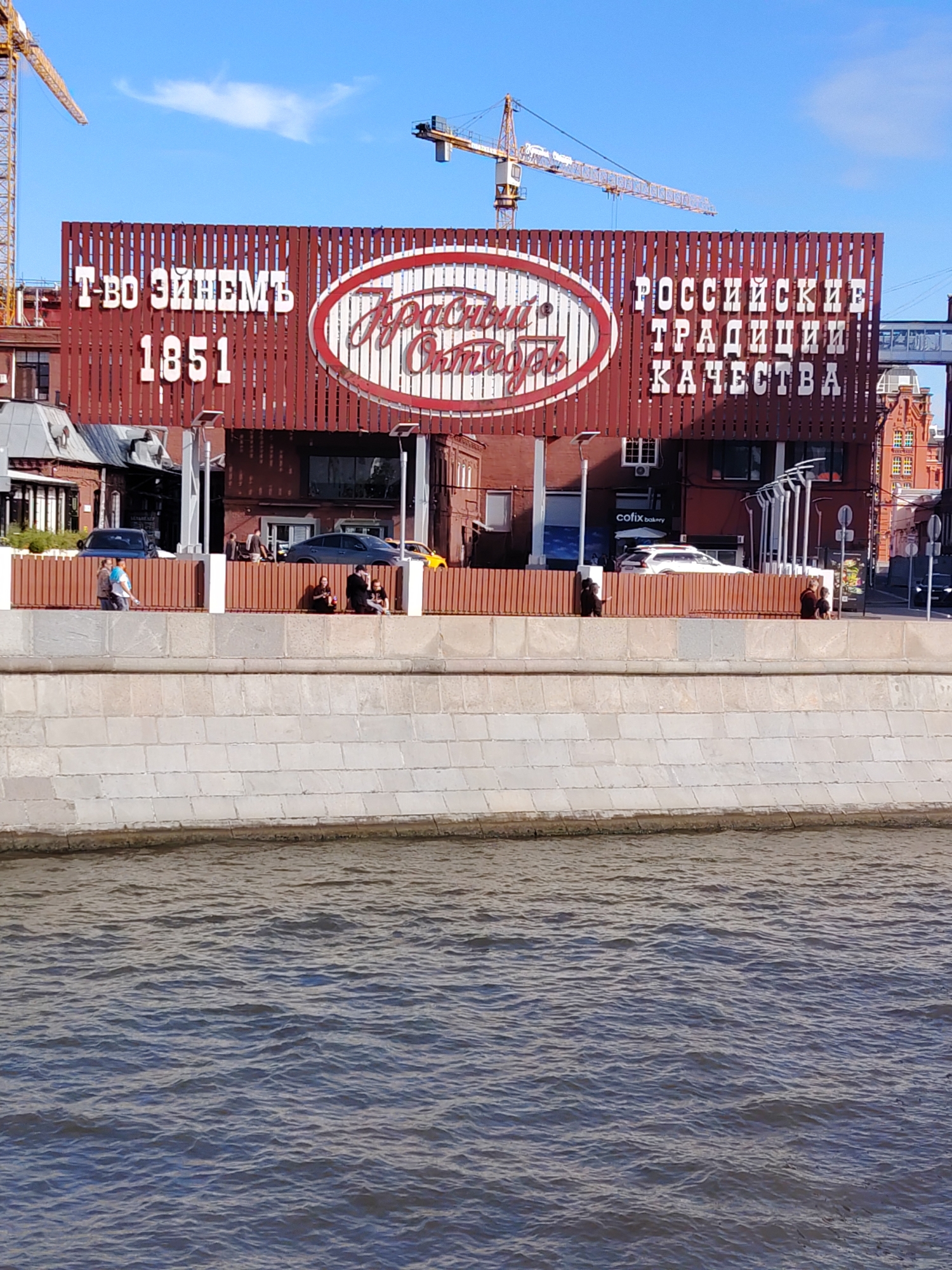 The image shows a large, dark red sign with white text and a red and white oval logo. The sign is mounted on a building with a wooden slat facade. The text on the left side reads "Т-ВО ЭиНЕМЬ" and "1851". The oval logo contains what appears to be handwritten text. The text on the right side reads "РОССИЙСКИЕ ТРАДИЦИИ КАЧЕСТВА". Several cranes are visible in the background, suggesting construction. In front of the building, there is a light-colored concrete embankment and a body of water. A few people are visible on the embankment, and some cars are parked in front of the building. The sky is blue with some white clouds.