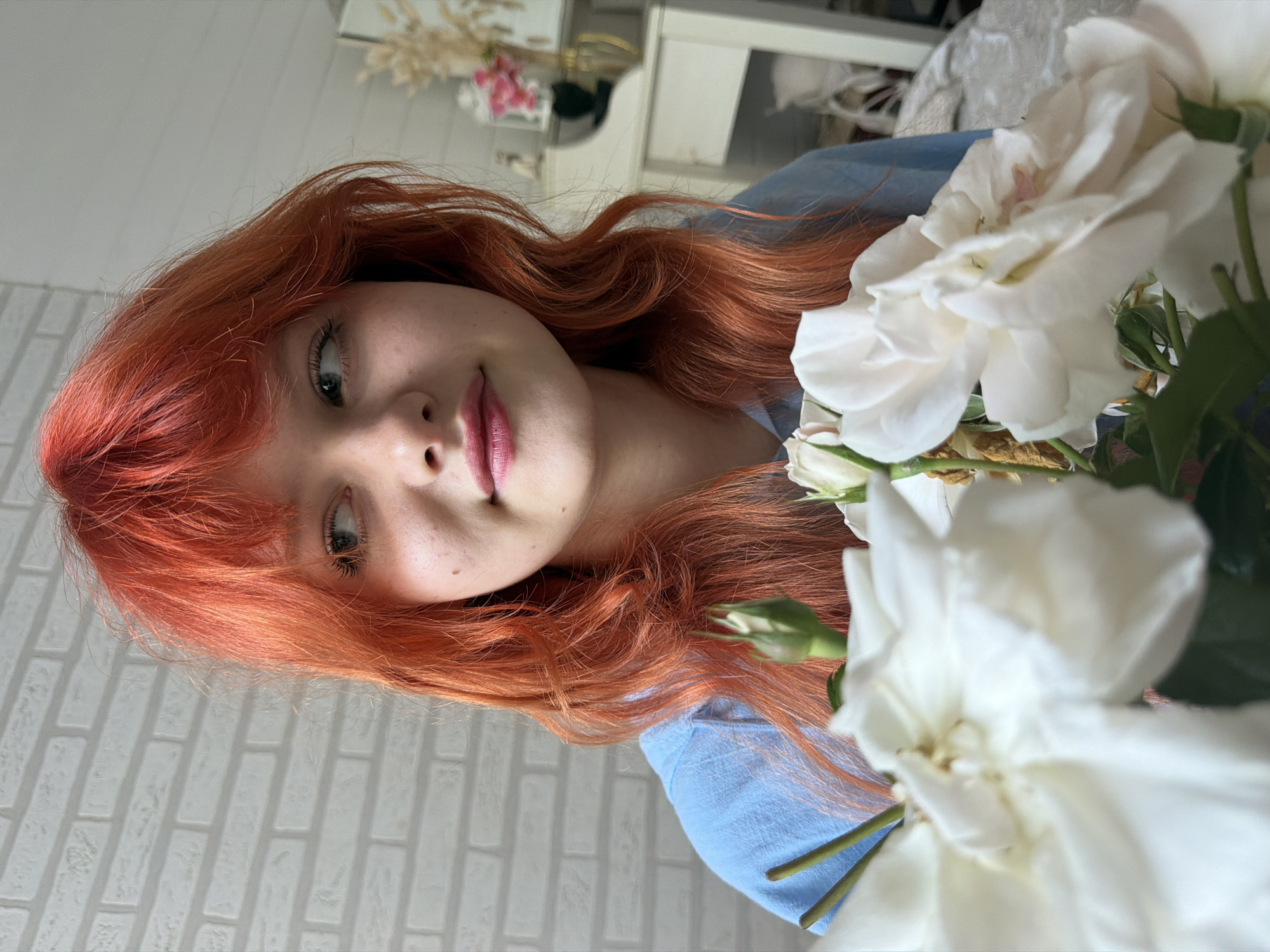 The media file is a vertical, close-up, eye-level shot of a young woman with vibrant, long, reddish-orange hair. She is looking slightly upwards and to her left, with a gentle smile playing on her lips. Her makeup is subtle, with pink lipstick and mascara accentuating her features. She is wearing a light blue garment, possibly a shirt or dress.
In the foreground and to the right of the woman, there are several white roses in bloom, some fully open and others as buds. Their delicate petals are soft and luminous. The background is blurred but suggests an indoor setting with a light-colored, textured wall that resembles white brickwork. Further in the background, a reflection in a mirror shows what appears to be a dresser or vanity with some decorative items, including dried grasses and pink flowers, hinting at a domestic or personal space.
The lighting suggests it might be daytime, with soft, diffused light illuminating the scene. The overall mood conveyed is one of gentle beauty and tranquility. There is no visible text in the media file. The location context provided is Sochi, Russia.