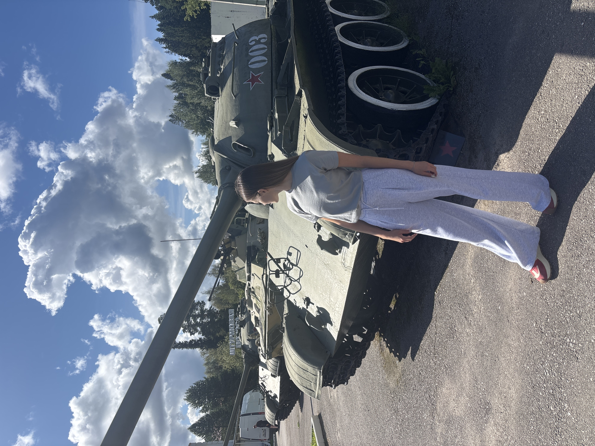 A young woman stands in front of a military tank outdoors on a sunny day. The tank is green with white markings, including the number "003" and a red star. The woman is wearing a grey t-shirt and grey sweatpants, with red and white sneakers. Her hair is long and brown, and she is looking to her right, away from the camera. The ground is paved, and there are trees and a blue sky with white clouds in the background. There is another military tank visible behind the one in the foreground. The location is identified as the city of Gamy, Russia.