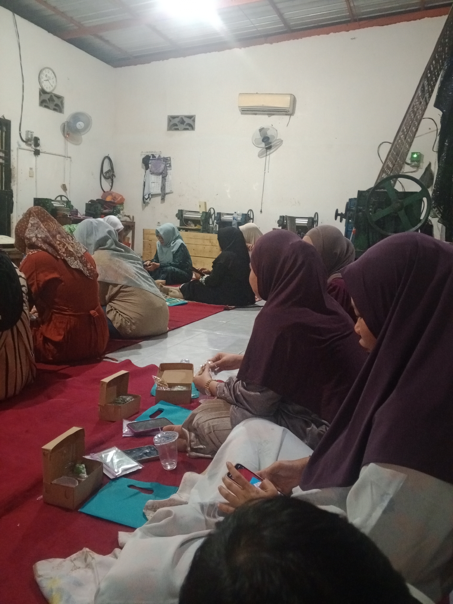 A group of individuals, mostly women wearing hijabs, are gathered on a red carpeted floor in what appears to be a workshop or production area in Mlilir, Indonesia. The room has a corrugated metal ceiling and white walls. Various items are scattered around, including small cardboard boxes containing what seem to be finished products, possibly jewelry or crafts, along with plastic packaging and turquoise tote bags. There are also cell phones and a clear plastic cup on the floor. In the background, industrial machinery, including what looks like a large green wheel and a metal press, is visible, suggesting a manufacturing environment. A clock and a fan are mounted on the wall. The lighting is dim and appears to come from overhead fluorescent lights. The individuals are seated and engaged in an activity, with some looking at their phones and others interacting with the small boxes. One person in the foreground is holding a smartphone. The scene suggests a collective work session or a distribution of goods in a modest industrial setting in Mlilir.