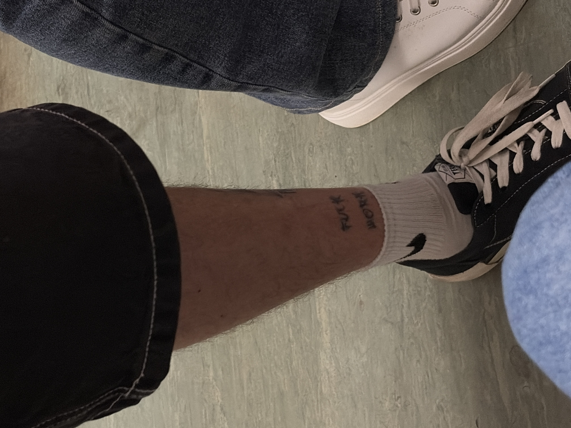 The image, specified as depicting a scene in Moscow, Russia, presents an overhead view of the lower legs and footwear of at least two individuals on a light-colored, possibly textured floor.
On the left, a person's bare lower leg is visible, clad in black shorts with a white stitched hem. The leg has visible hair. A tattoo is present on the lower shin/upper ankle area, appearing to read "FUCK" on the upper line and "WORK" on the lower line. A white ankle sock with a black Nike swoosh logo on the outer side covers the foot, which is inserted into a dark-colored sneaker with white laces and a white sole. A faint light blue fabric, likely clothing, is partially visible at the bottom left of the frame, possibly belonging to a third individual or the same individual wearing the sneaker.
On the right, the lower leg of another person is visible, covered by blue denim jeans. This individual wears a white sneaker with a thick white sole and visible eyelets for laces, though the laces themselves are not clearly detailed. The two individuals are in close proximity, suggesting they are seated or standing near each other. The floor surface exhibits subtle variations in tone, consistent with a worn or patterned indoor flooring material.