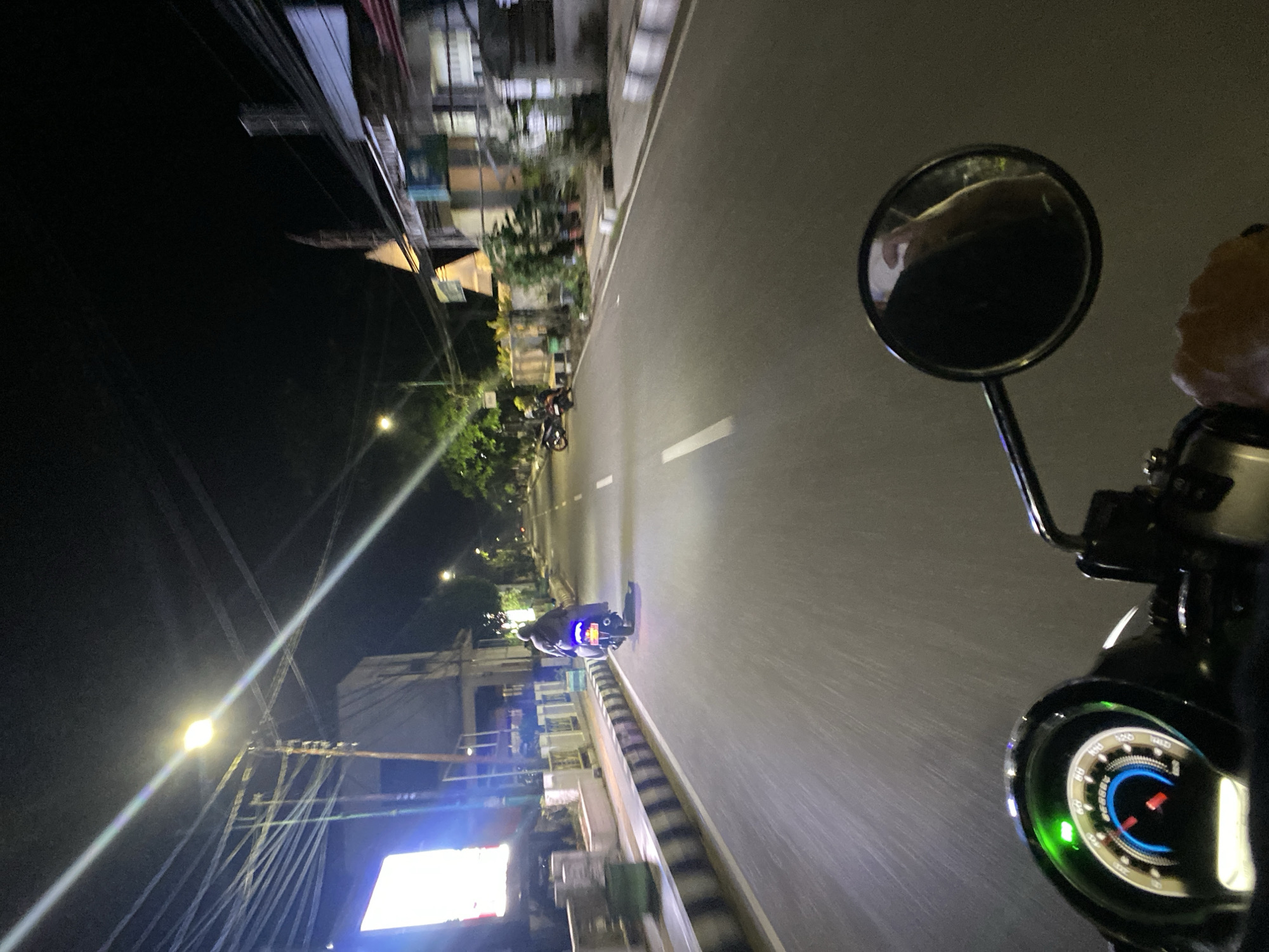 The image depicts a night scene on a road in Tanjung, Indonesia, taken from the perspective of a person riding a motorcycle. Several other motorcycles are visible further down the road, traveling in the same direction. The road is illuminated by streetlights, and buildings line both sides. A motorcycle mirror is visible on the right side of the frame, and part of the rider's hand can be seen. The dashboard of the motorcycle is also partially visible. Electrical wires crisscross above the road.