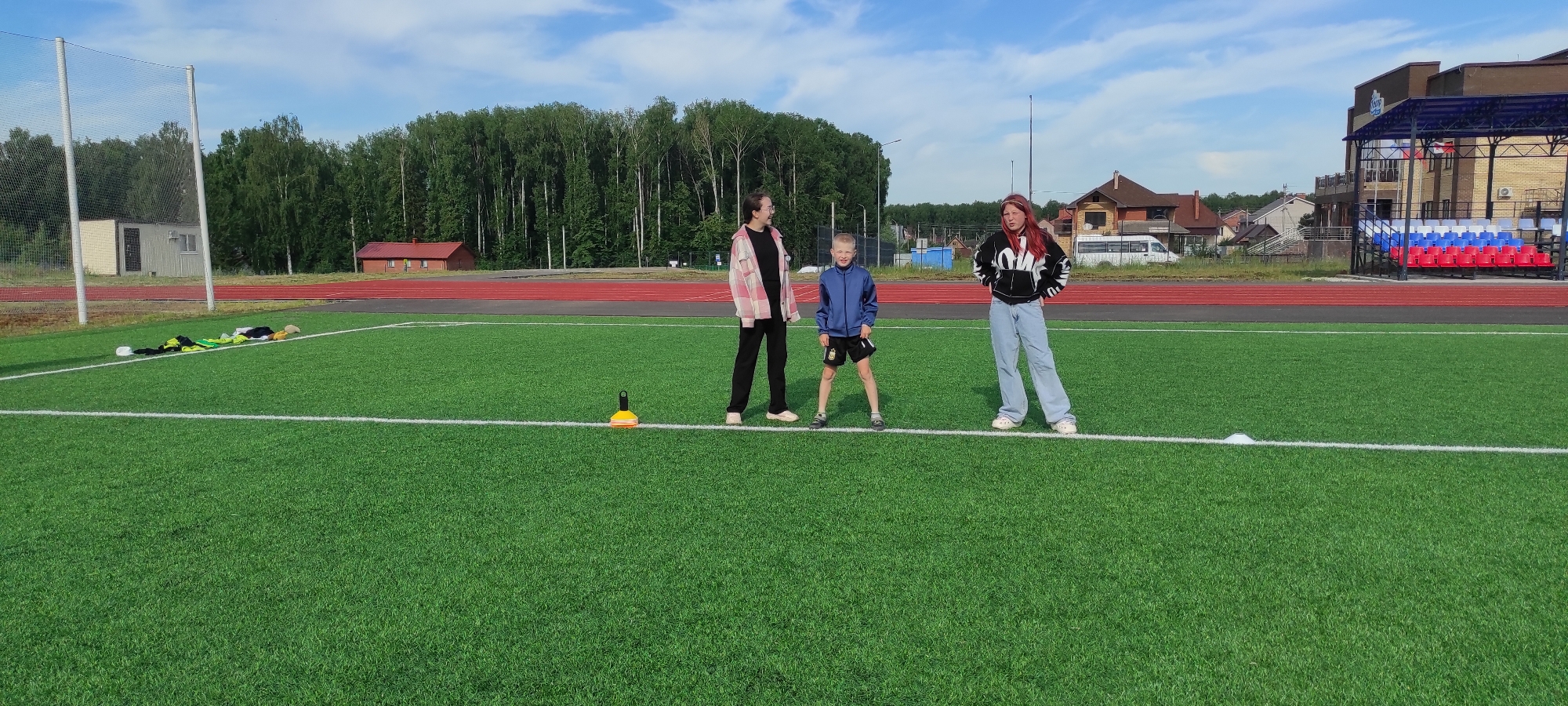 The image shows an outdoor athletic field, likely a soccer field, with bright green turf and white boundary lines. In the foreground, on the turf, are several pieces of athletic apparel, including a yellow and green jersey, black shorts, white shorts, and a tan cap. There is also a yellow cone on the field.
In the mid-ground, three young people are standing on the turf. From left to right, the first person is a young female wearing a black t-shirt and a pink and white plaid jacket, with black pants and light-colored shoes. She is looking to her right. The second person is a young male wearing a blue jacket, black shorts with white trim, and dark shoes. He is looking forward. The third person is a young female wearing a black hooded sweatshirt with a white design, light blue jeans, and white shoes. She has red hair tied back and is looking to her left with her hands on her hips.
In the background, there is a red track surrounding the field. Beyond the track, there is a dense line of green trees. To the left, there is a small, beige building with a white door and window. To the right, there is a modern stadium with a blue and silver roof and rows of red, white, and blue seats. Several houses are visible behind the stadium. The sky is blue with scattered white clouds.
The location is identified as an athletic facility in Noviy, Russia, based on the provided information.