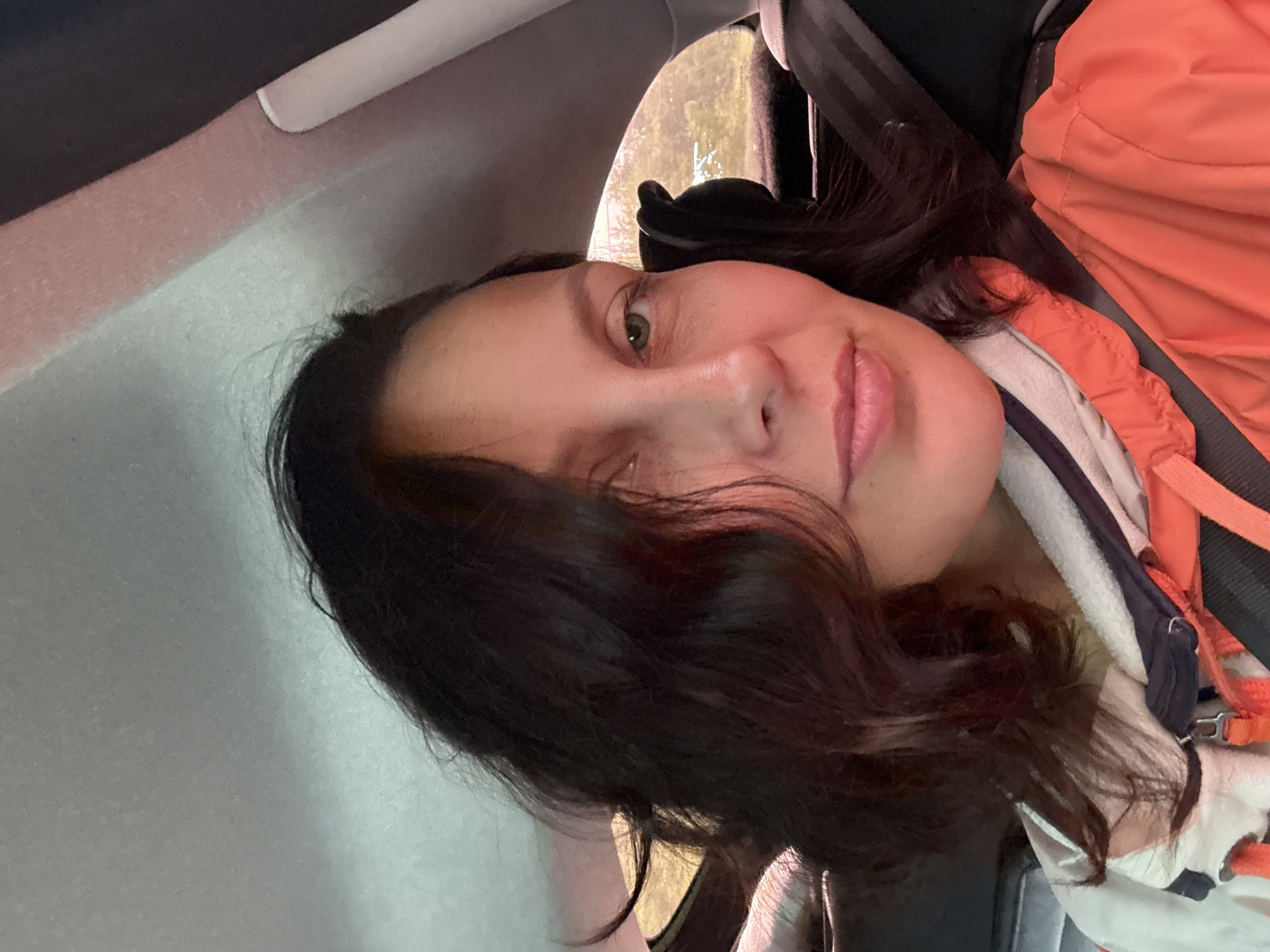 A close-up, selfie-style, medium shot captures a woman in the backseat of a vehicle. She has dark, wavy hair that falls around her face and shoulders. Her gaze is directed slightly away from the camera, with her left eye, a green hue, being more visible. Her lips are slightly parted and appear to be a pale pink. She is wearing an orange jacket over a white and dark-striped garment. A black seatbelt is visible across her chest and shoulder. The background shows the interior of a car, with a gray headliner and a window revealing a blurred outdoor scene with trees. The specific location is within the city of Gremyachinsk, Russia.