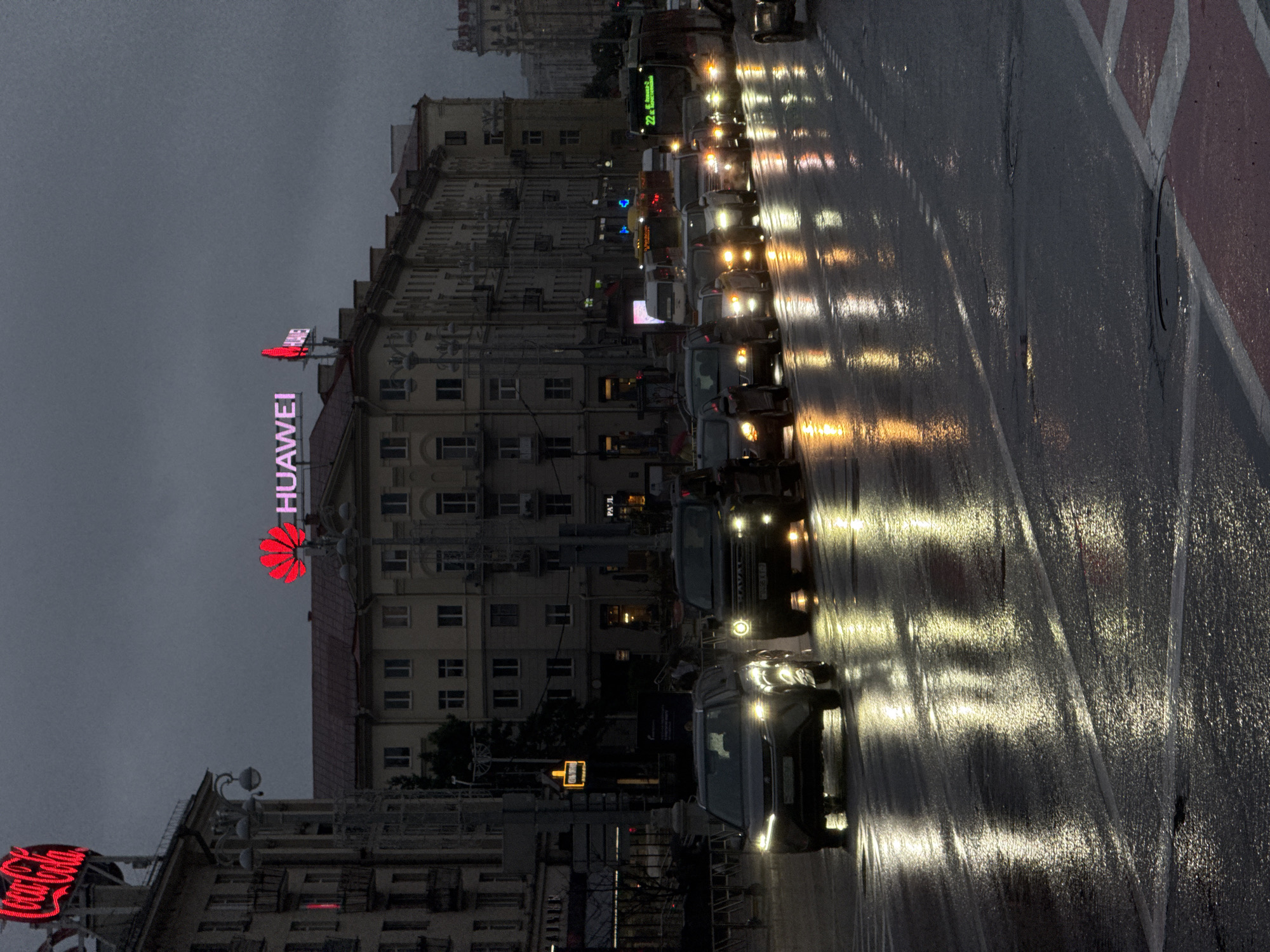 The image depicts a street scene in Minsk, Belarus, under a gloomy, overcast sky, likely during evening or twilight. The road is wet, reflecting the lights of vehicles and streetlamps, suggesting recent rainfall. A line of cars is visible, with headlights illuminating the wet asphalt. In the background, buildings line the street, some with illuminated signs. Notably, a large red neon sign reading "Coca-Cola" is visible on the left, and a prominent red Huawei logo with the company name in pink neon is displayed on a building further down the street. The overall atmosphere is urban and somewhat somber due to the weather and lighting conditions. There are no visible people in the immediate foreground, though the presence of traffic implies human activity. The scene appears to be a typical city street during inclement weather.
