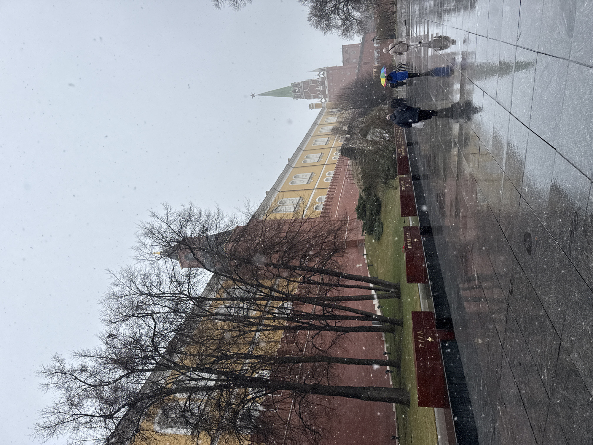 The image shows a snow-covered scene in Moscow, Russia, with a view of the Kremlin wall and one of its towers. Bare trees stand in front of the red brick wall and a light-colored building section. Several people are walking along the wet stone pavement beside the wall, one holding a colorful umbrella. Small memorial monuments are situated between the trees and the path. Snowflakes are visible in the air.